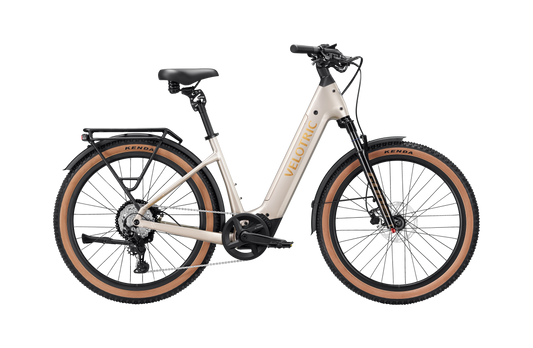 A cream-colored Velotric Discover M comfort eBike by Velotric, featuring a step-through frame, black seat, rear rack, front and rear fenders, and tan tires is shown in profile against a white background.