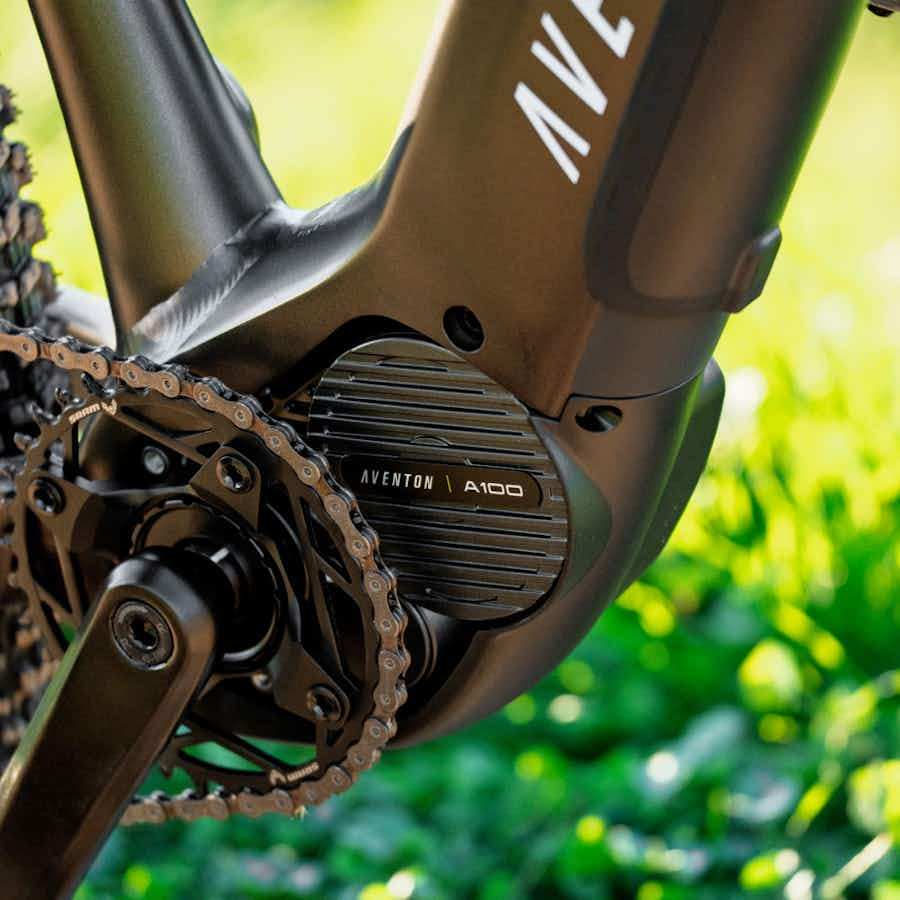 Close-up of the Aventon Ramblas ADV electric mountain bike’s mid-drive motor and crankset by Aventon, with its frame and green grass in the background.