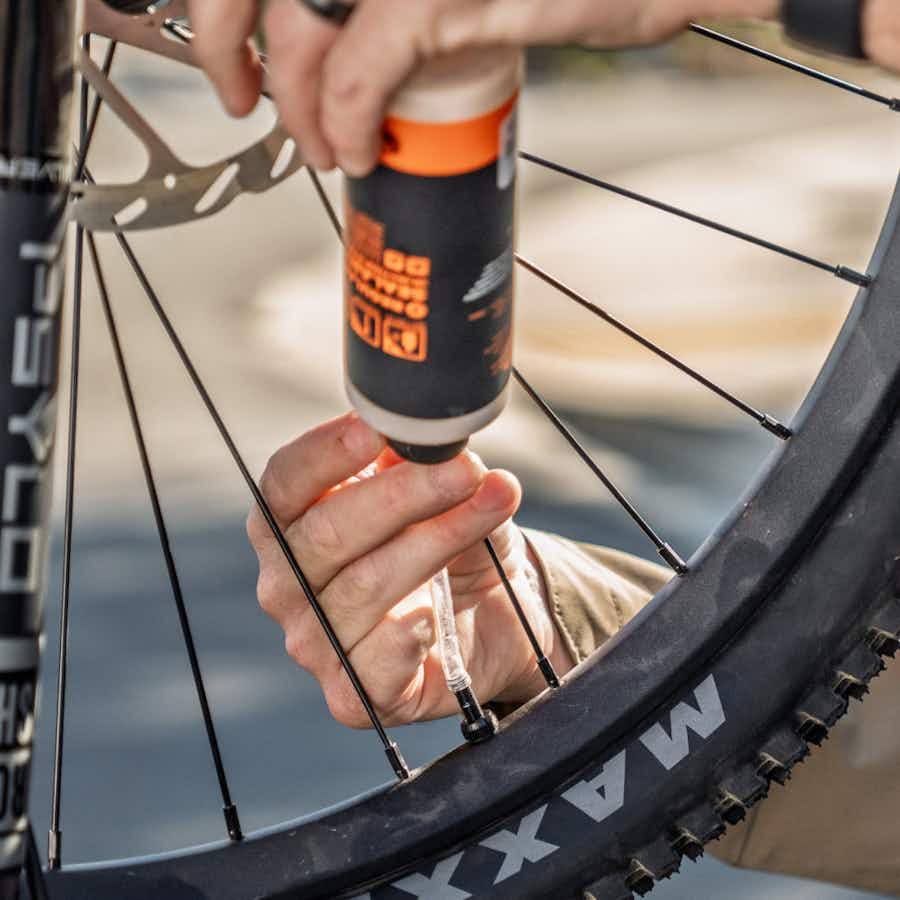 A person is injecting sealant into a bicycle tire through the valve stem while steadying the Aventon Ramblas ADV electric mountain bike from Aventon.