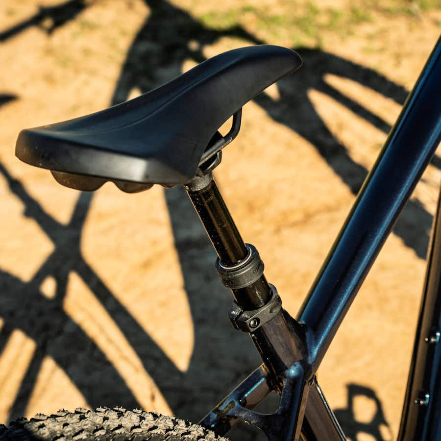 Close-up of a black seat and seatpost on an Aventon Ramblas ADV electric mountain bike by Aventon, with part of the wheel and ground visible in the background.