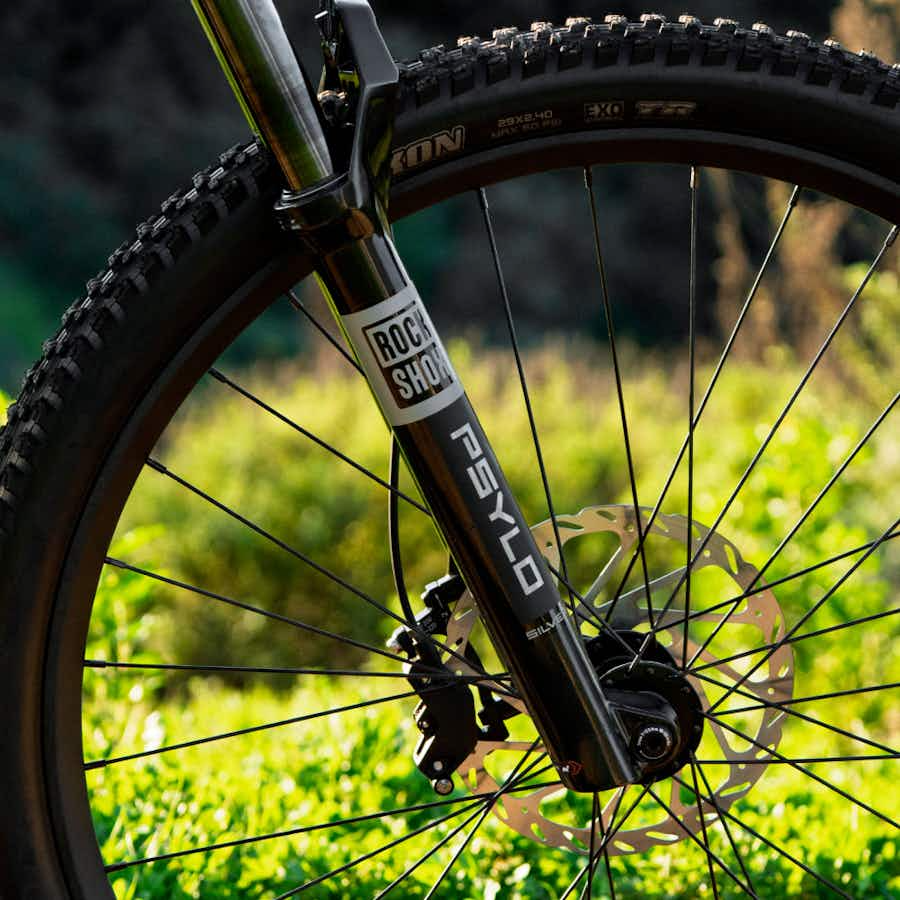Close-up of the Aventon Ramblas ADV electric mountain bike's front wheel, featuring a RockShox Psylo suspension fork, disc brake, and knobby tire by Aventon, set against a blurred outdoor background.