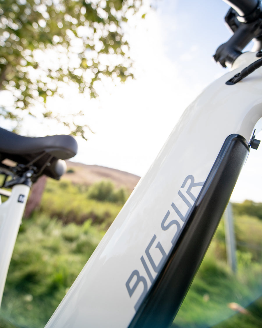 Close-up of a sleek white bicycle frame with "AIMA - Big Sur" elegantly displayed, set against a blurred outdoor backdrop of trees and sky. This all-terrain marvel from AIMA is equipped with a powerful motor, ready for any adventure nature throws your way.