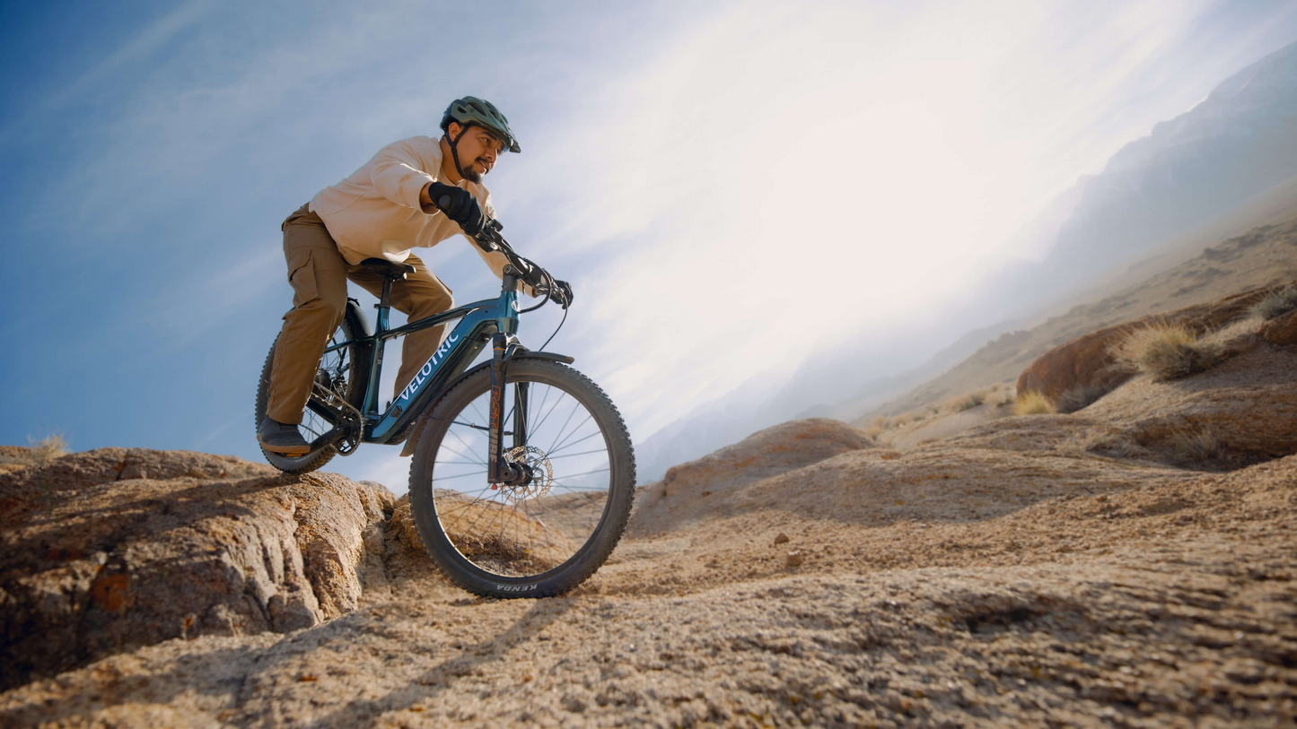A rider in a helmet and outdoor gear speeds down a rocky trail on the Velotric Summit 2 hybrid eBike by Velotric, set against a dry, open landscape and bright sky.