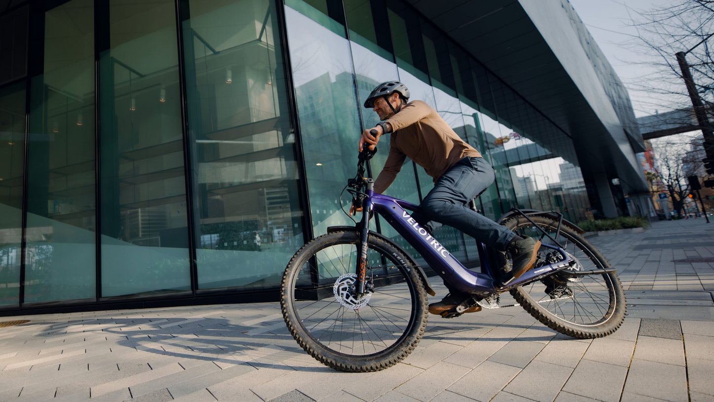 A person in a helmet rides a blue Velotric Summit 2 electric bike by Velotric along a paved sidewalk beside a modern glass building.