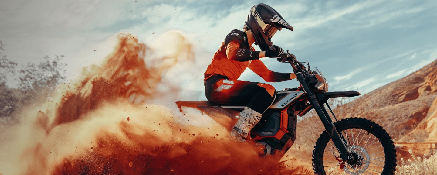 Riding through sandy terrain, a motorcyclist in orange gear kicks up red-brown dust on a Segway Xaber 300 electric dirt bike by Segway, with mountains visible in the background.