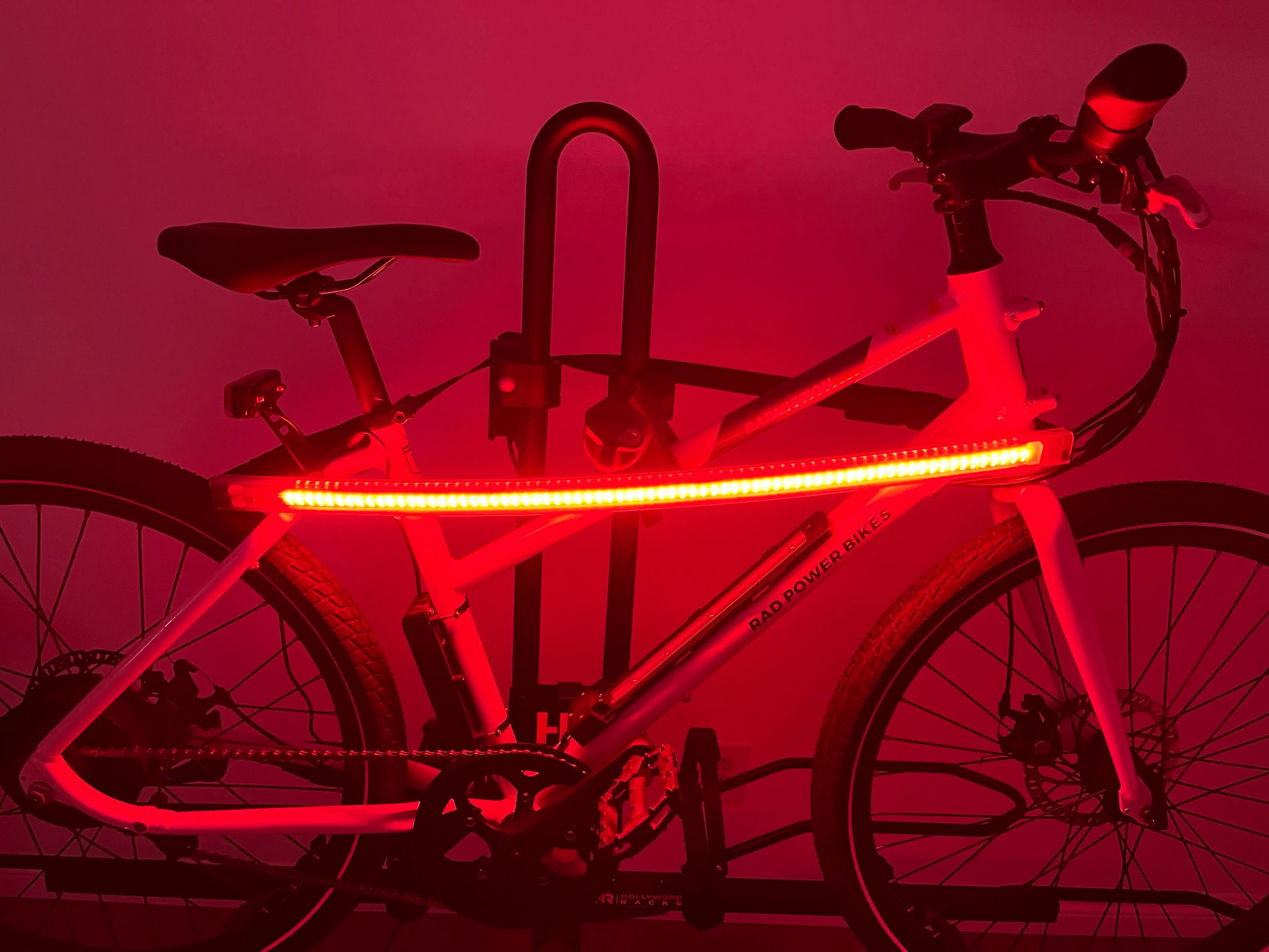 A white electric bike with a Hollywood Racks - Safety Light Bar glowing red and secured on a bike rack in a dark setting. The words "RAD POWER BIKES" are visible on the bike frame.