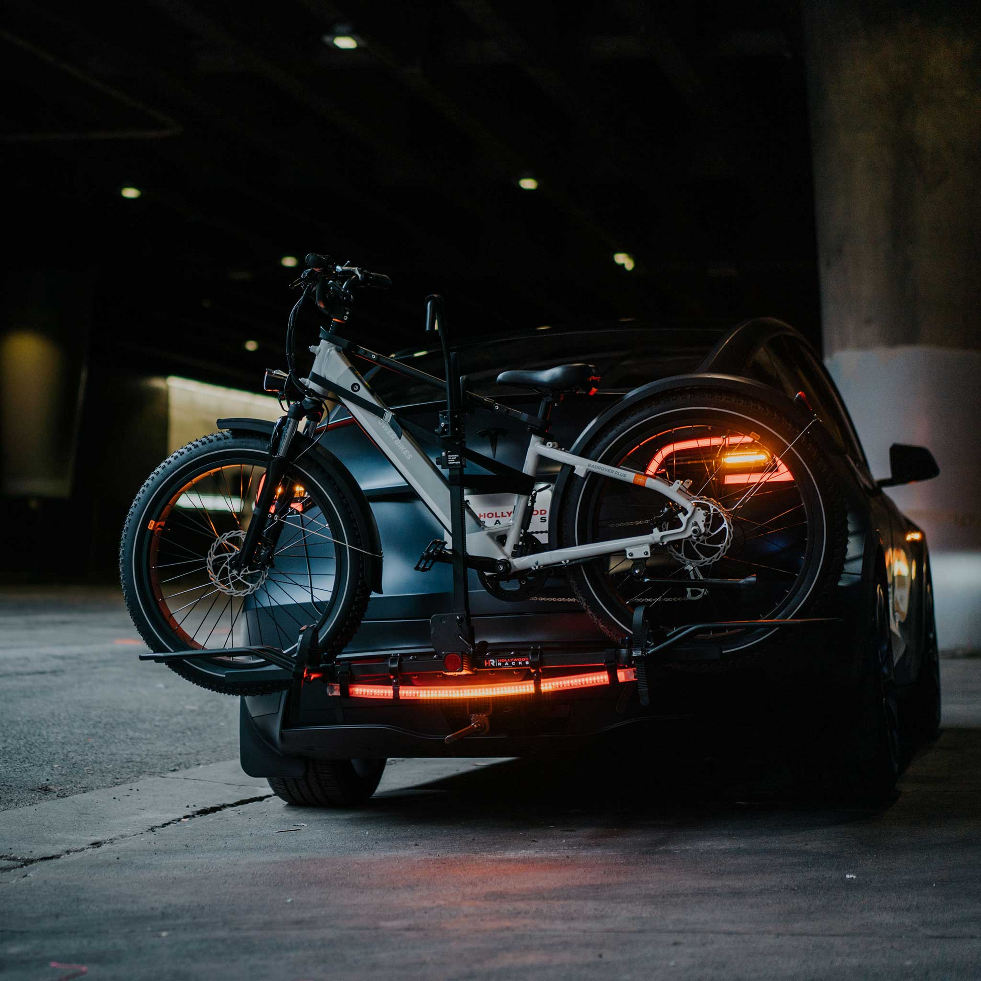 In a dimly lit parking area, a bicycle is secured on the rear rack of a car, with the Hollywood Racks - Safety Light Bar and the bike's reflectors both illuminated.