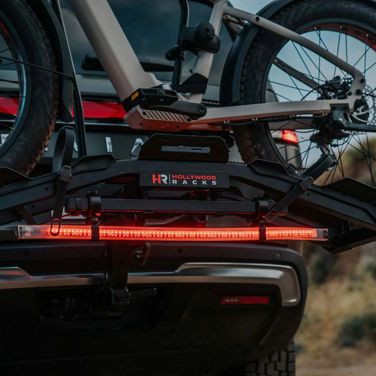 A Hollywood Racks bike carrier, securely mounted on the rear of an SUV with two bicycles attached, features the Hollywood Racks Safety Light Bar Adapter for enhanced visibility and bike safety. The setup is set against an outdoor backdrop.