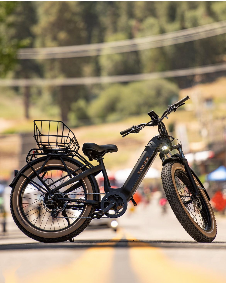 The AIMA - Big Sur eBike, renowned for its powerful motor and all-terrain thick tires, is parked on a road. It includes a front basket and rests against a blurred backdrop of trees and power lines.