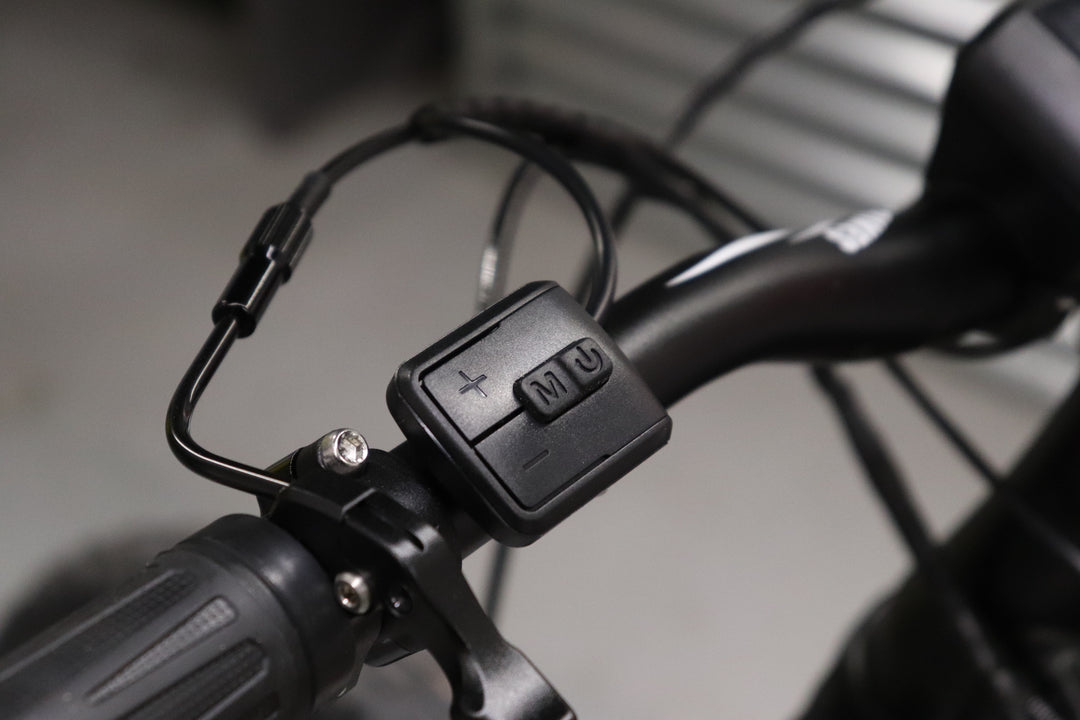 Close-up of a bicycle handlebar showing control buttons for pedal assistance. The buttons are labeled with plus, minus, and a middle button marked with an "M." This setup is often found on LMX - 64 by LMX featuring a brushless mid-mounted motor.