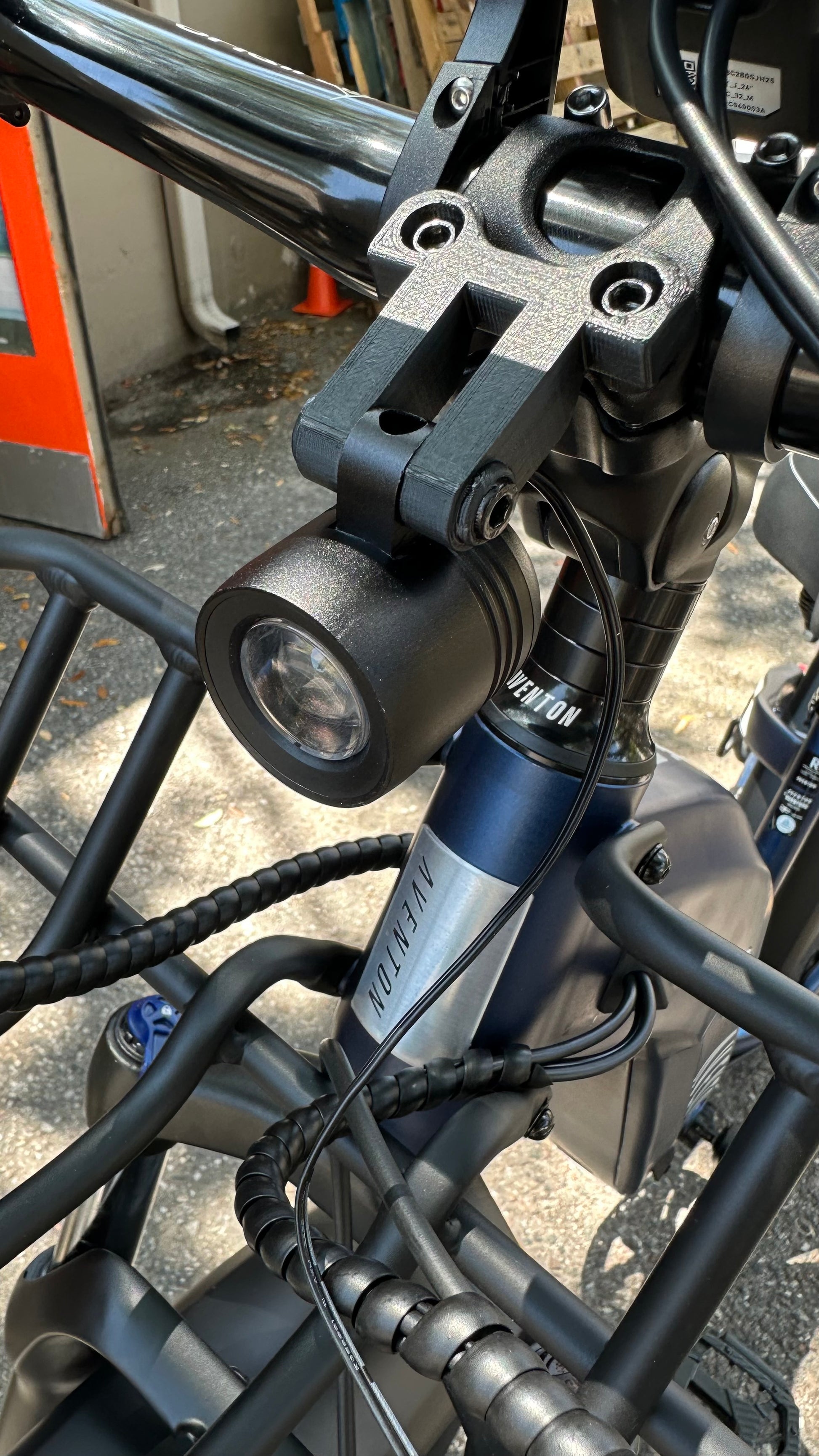 Close-up view of an eBike accessory—a bicycle front light mounted on the handlebars, with wires and parts of the bicycle frame visible. The Tampa Bay eBikes Universal Adjustable Stem Face Plate Accessory Mount secures the light efficiently, enhancing its stability.