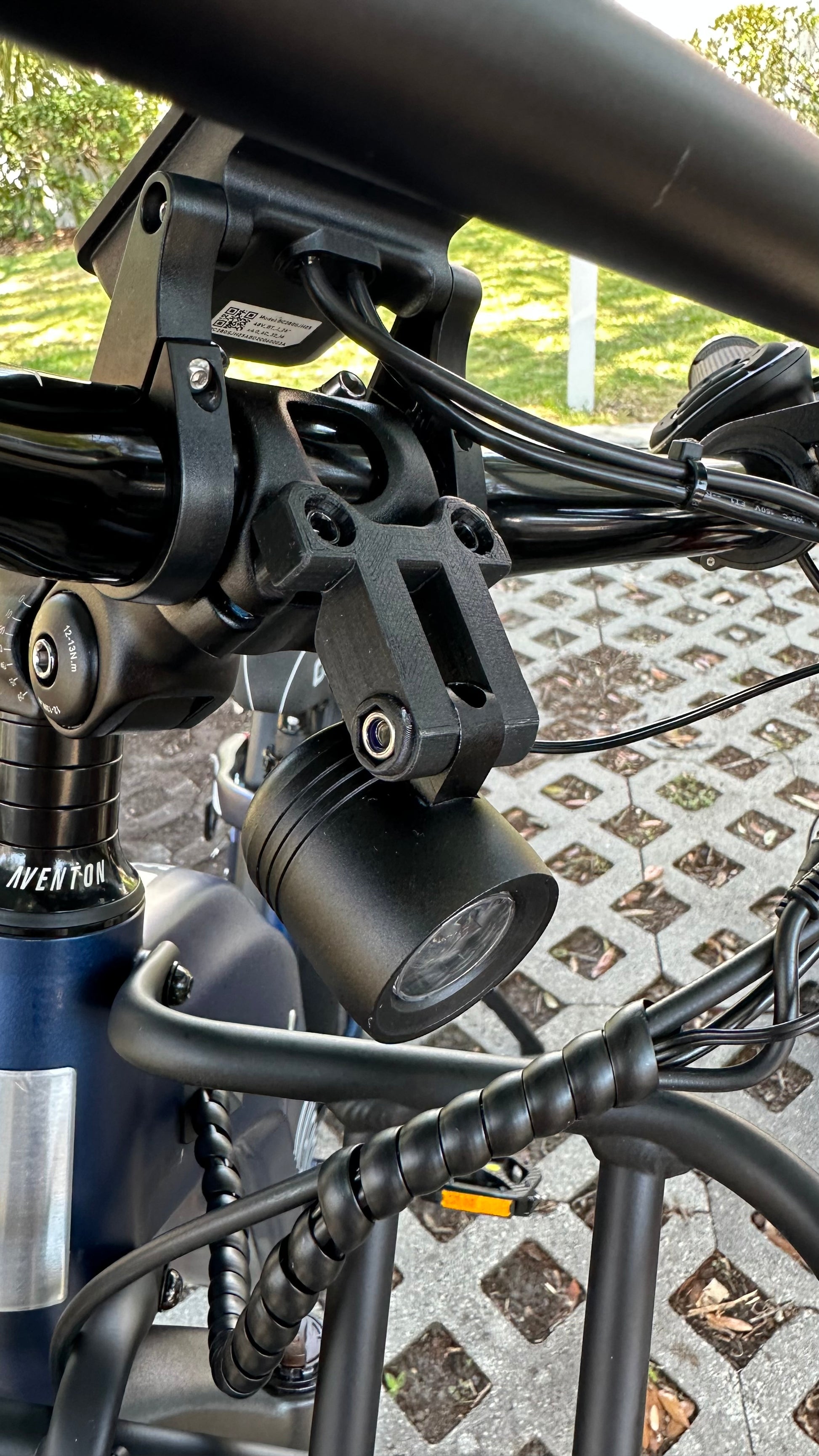 Close-up of a bicycle handlebar equipped with the Tampa Bay eBikes Universal Adjustable Stem Face Plate Accessory Mount, featuring an eBike headlight. The background displays a paved area surrounded by greenery.