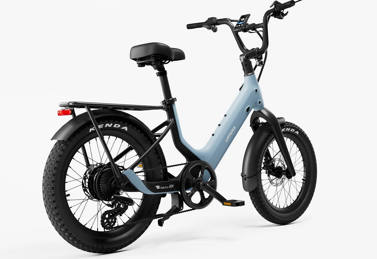 Urtopia Carbon Joy Ultra electric cruiser bicycle by Urtopia in light blue and black, featuring wide tires, rear cargo rack, front suspension, and a digital display, shown from a rear three-quarter angle on a plain white background.
