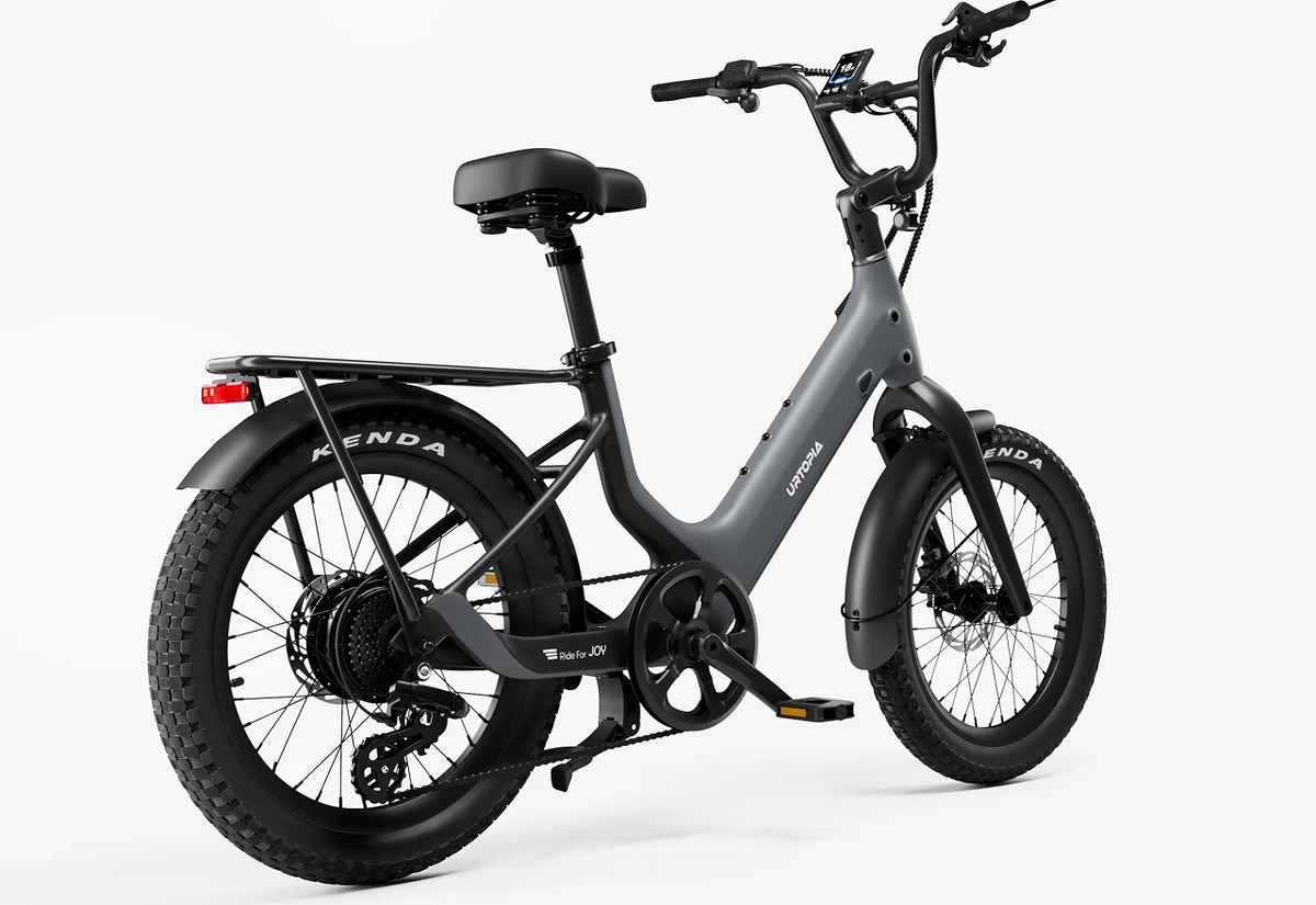 The Urtopia - Carbon Joy Ultra by Urtopia is a matte gray electric cruiser bicycle featuring wide tires, a rear cargo rack, black saddle, handlebar-mounted display, and a long-range battery, positioned at an angle on a white background.