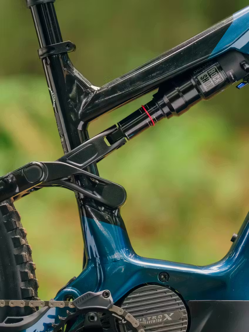 Close-up of the Aventon Current ADV full-suspension eMTB frame by Aventon, highlighting the rear suspension system, partial rear wheel, and RockShox shock absorber against a blurred outdoor background.