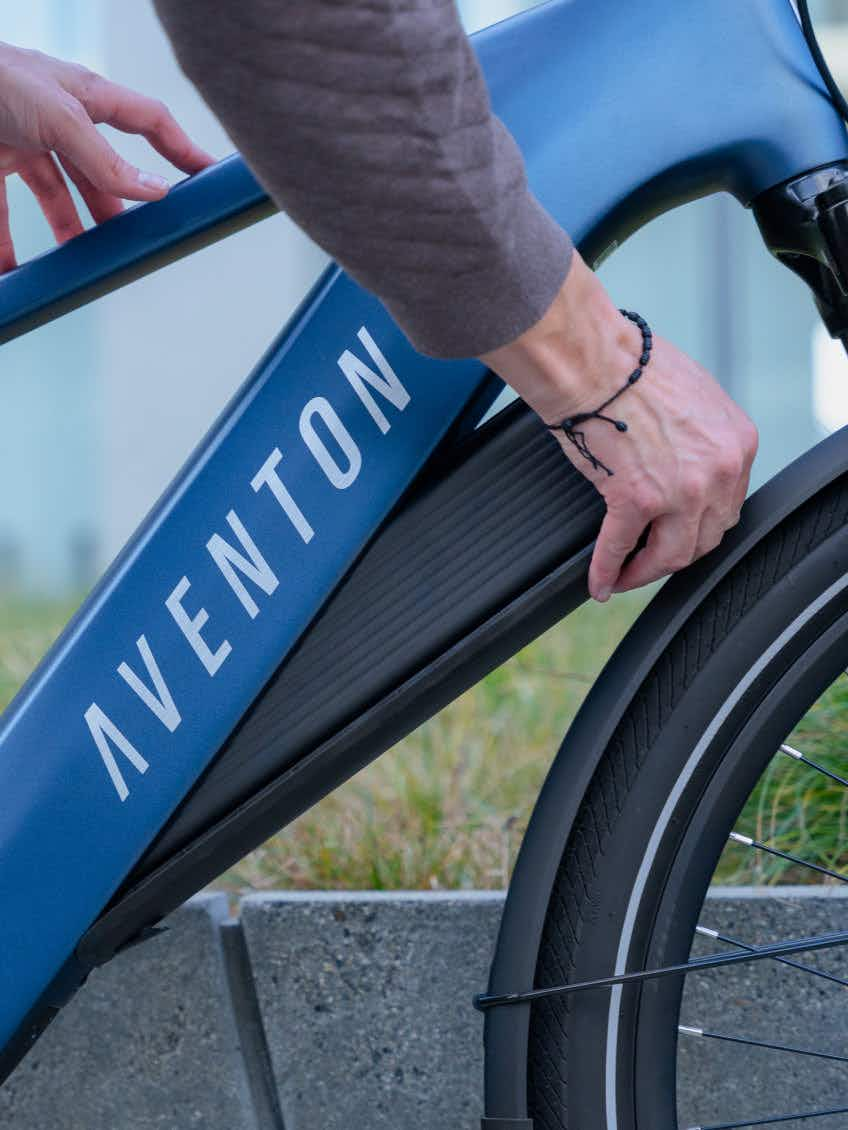 A person inserts or removes the battery from the frame of an Aventon Level 4 REC electric bike by Aventon, parked near a concrete edge.