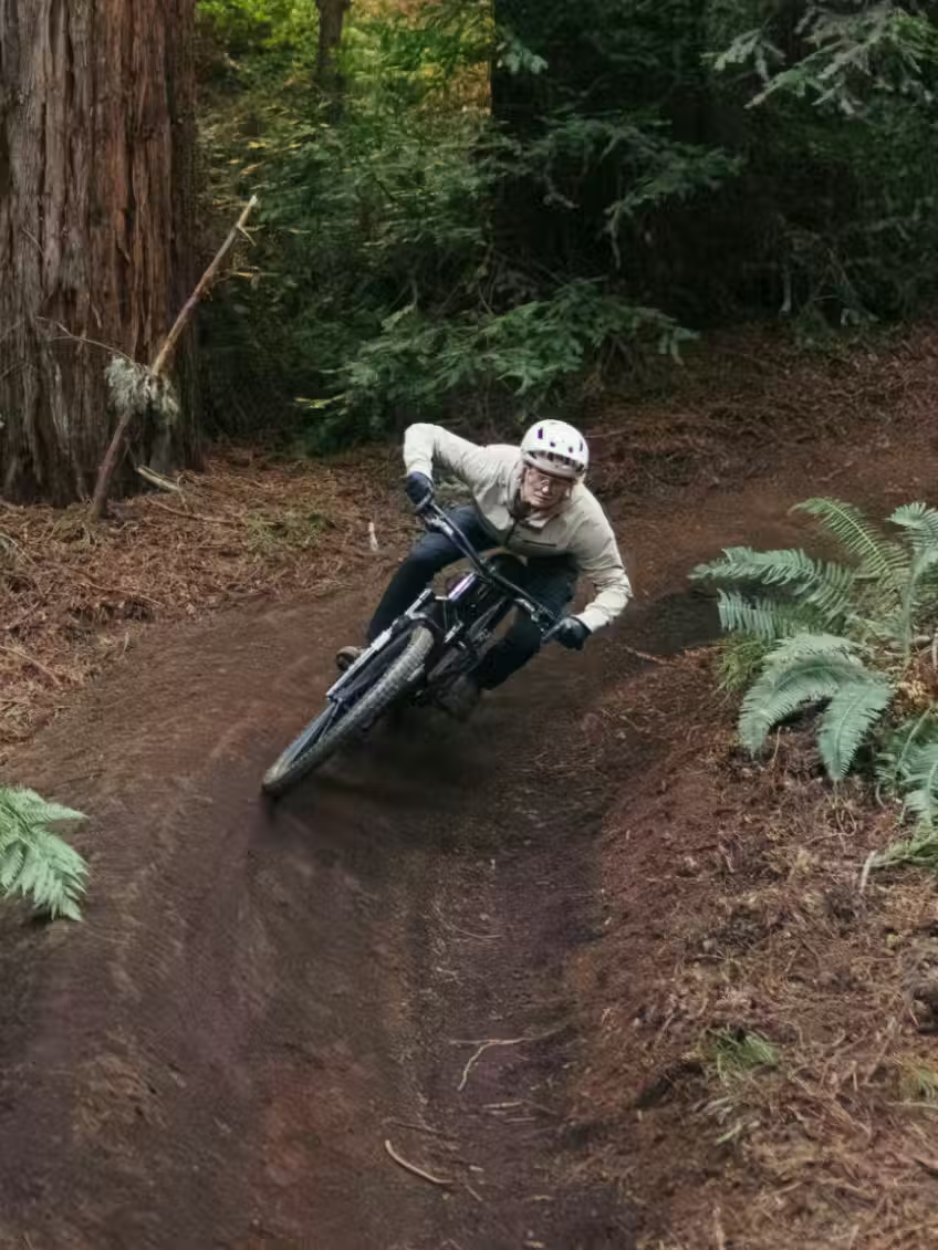A rider in a helmet navigates a dirt trail curve through ferns and tall trees on the Aventon Current ADV full-suspension eMTB by Aventon.