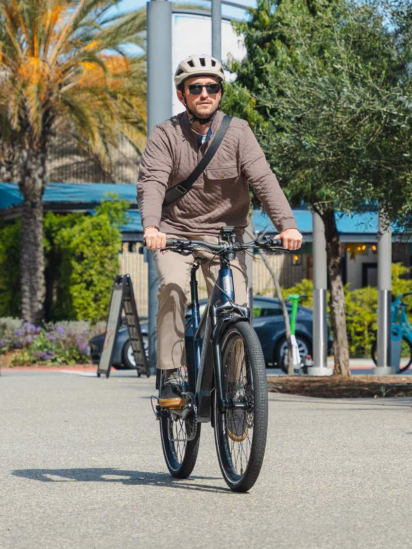 A man in a helmet and sunglasses rides an Aventon Level 4 REC electric bike from Aventon on a sunny day, with trees and parked cars in the background.
