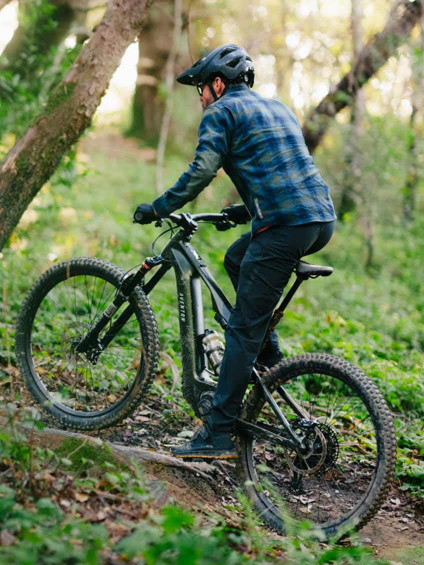 A rider in a helmet and plaid shirt pedals the Aventon Current EXP electric mountain bike by Aventon uphill on a dirt trail through a lush, wooded area.