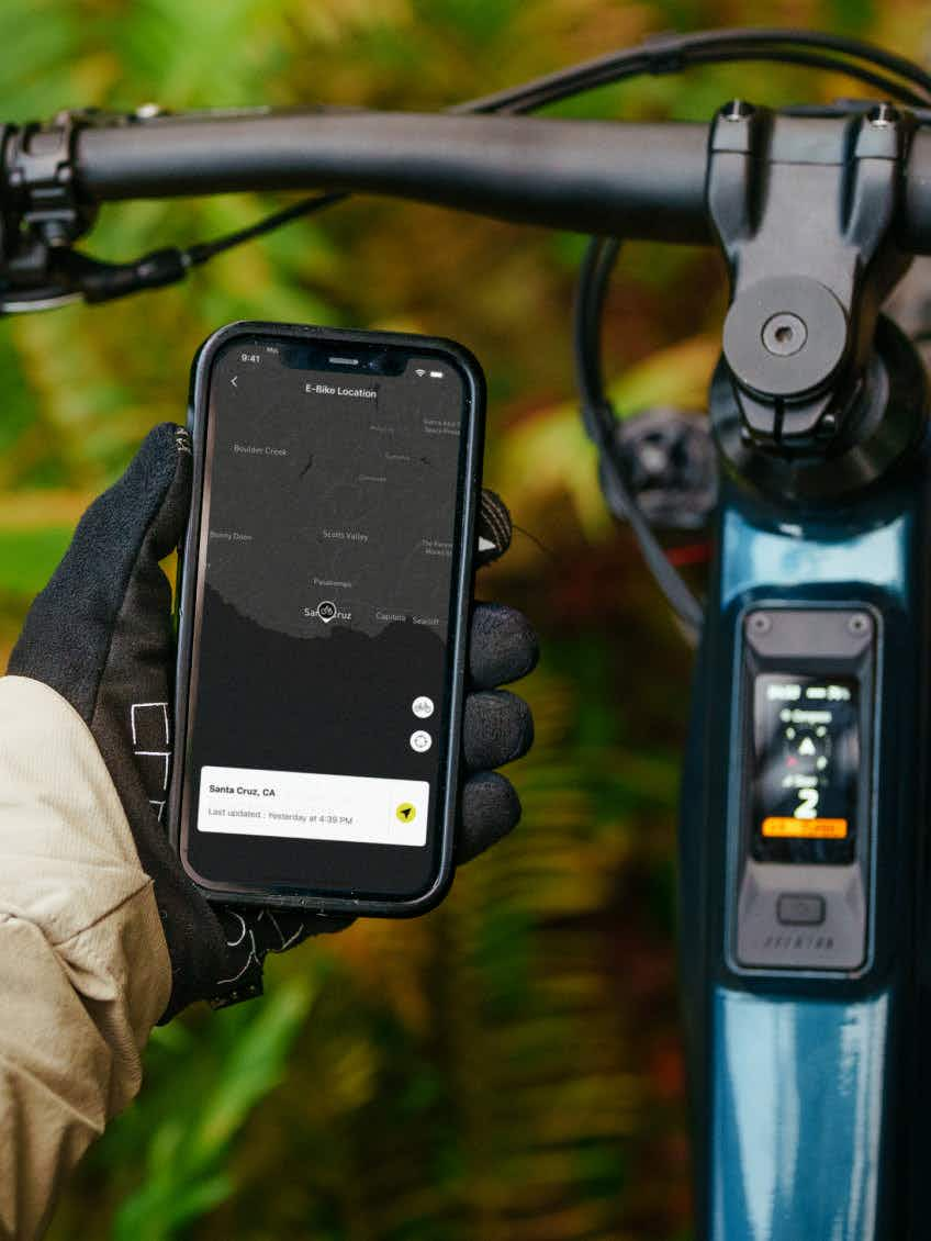 A gloved hand holds a smartphone showing an e-bike location app beside the handlebars and display of the Aventon Current EXP carbon eMTB by Aventon, set against a blurred outdoor background.