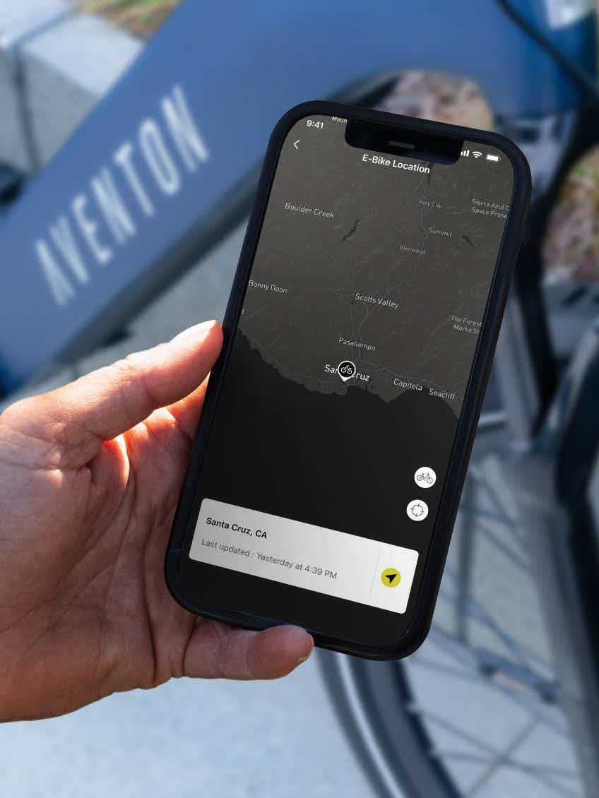 A hand holds a smartphone with an e-bike location map showing Santa Cruz, CA as the last update; in the background is an Aventon Level 4 REC electric bike by Aventon, featuring a powerful hub motor.
