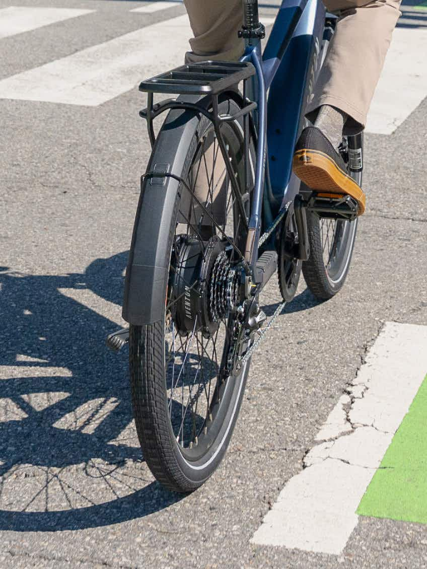 Close-up of a rider on the Aventon Level 4 REC by Aventon in an urban setting, featuring the rear wheel, gears, and part of their legs and shoes, all powered by a robust hub motor.