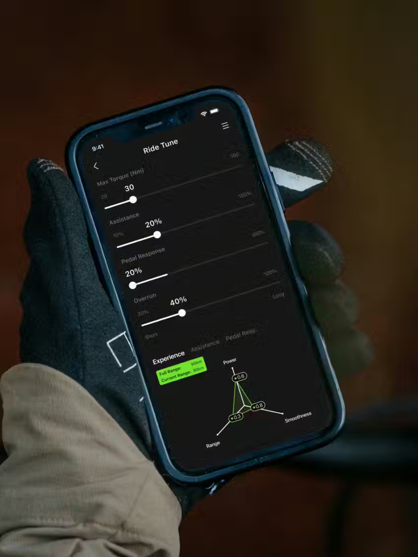 A gloved hand holds a smartphone showing the Aventon "Ride Tune" app with settings for max torque, assistance, pedal response, and overrun—perfect for customizing your Aventon Current ADV e-bike ride.