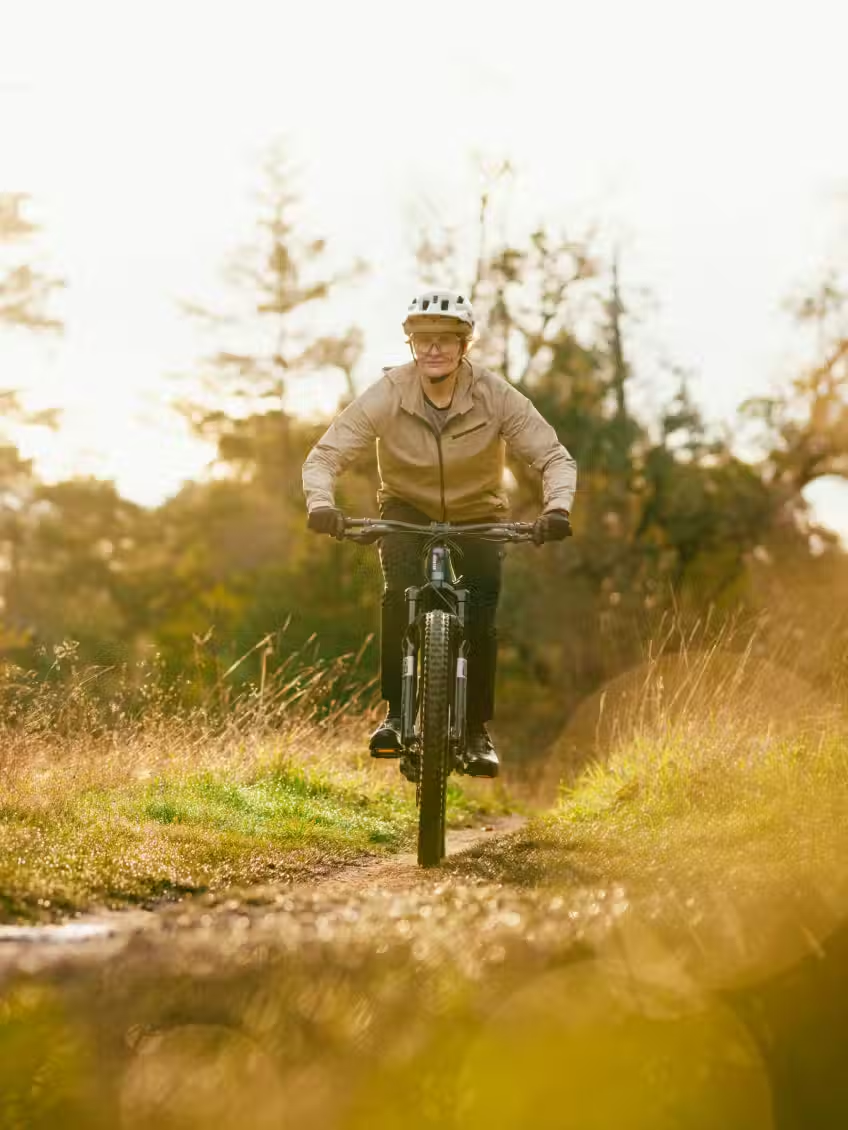 A rider in a helmet and jacket rides the Aventon Current ADV electric mountain bike by Aventon along a dirt path through grassy terrain with trees in the background.
