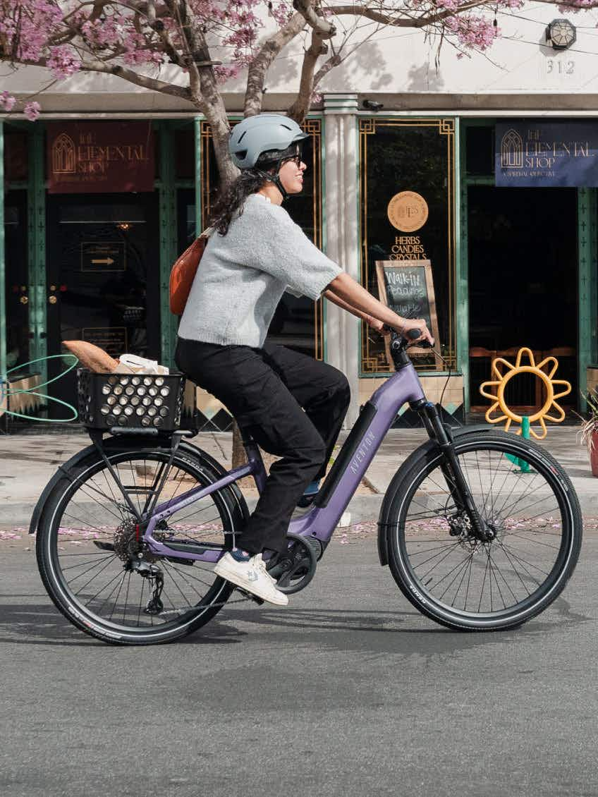 A helmeted rider cruises past a storefront and a flower-shaped bike rack on an Aventon Level 4 ADV commuter eBike in purple, with a basket carrying a baguette and groceries.