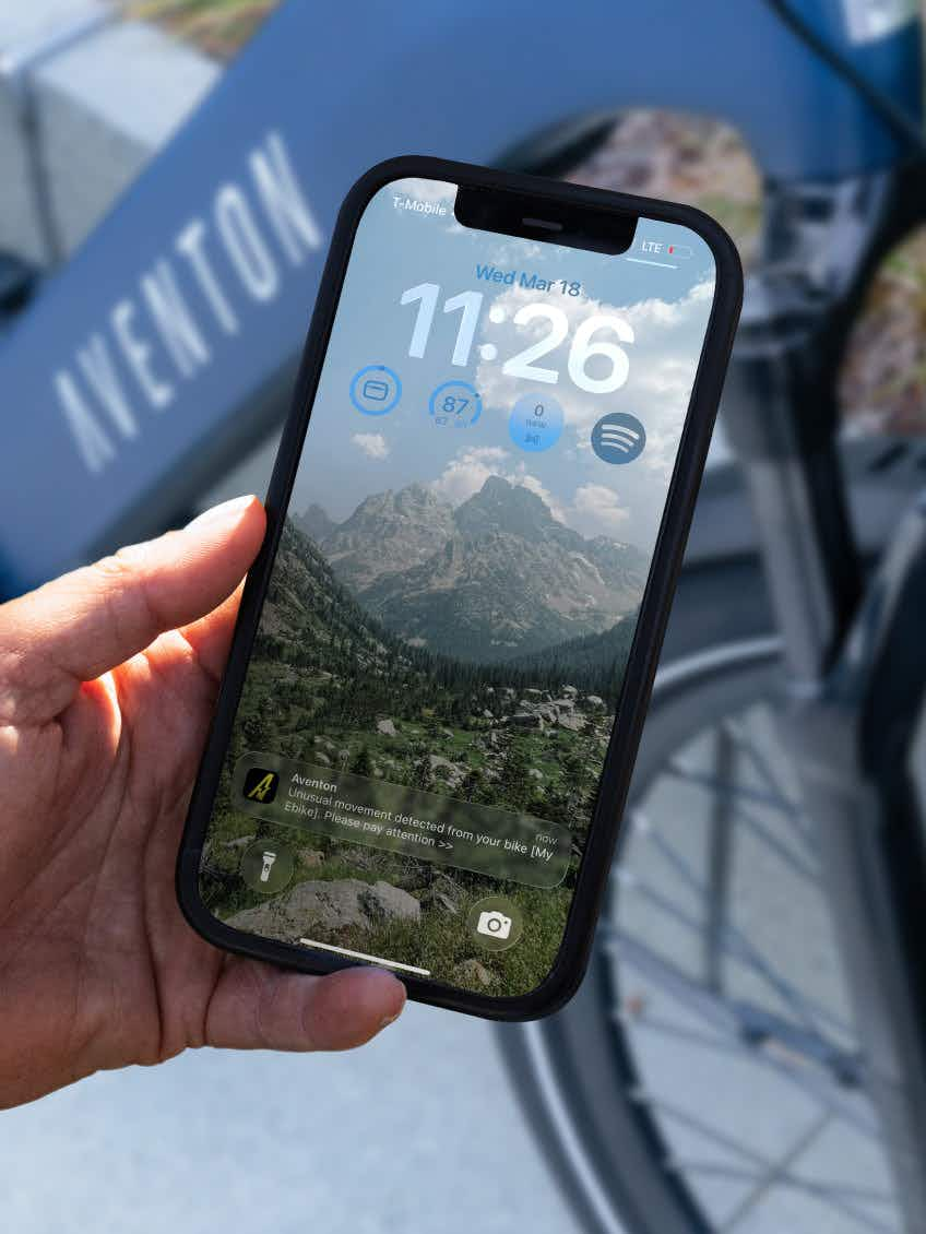 A hand holds a smartphone showing the lock screen with time, weather, and bike security alert in front of an Aventon Level 4 REC electric bike by Aventon, featuring a powerful hub motor and the “Aventon” logo on the frame.