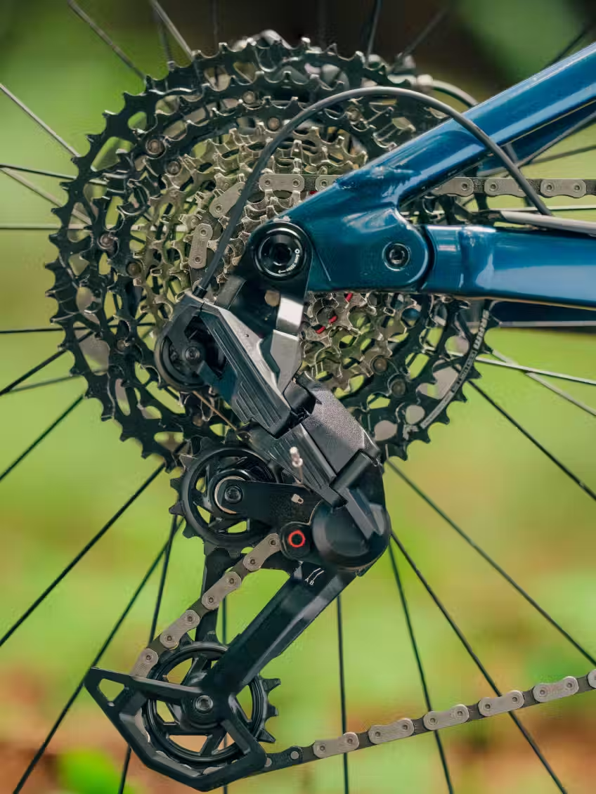 Close-up of the Aventon Current ADV by Aventon, showcasing the electric mountain bike’s rear derailleur, cassette, and chain with gears and components in focus against a blurred green background.