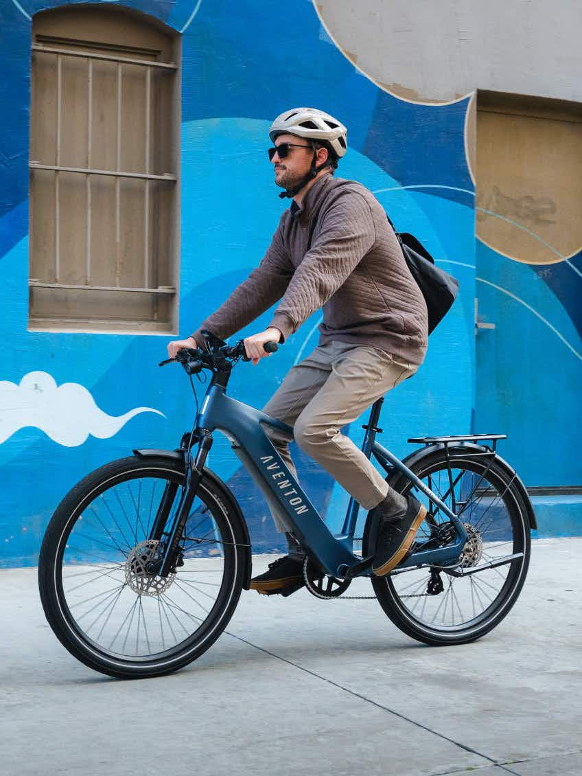 A rider in a helmet and sunglasses cruises past a blue mural on a city sidewalk on the Aventon Level 4 REC electric bike by Aventon, which features a powerful hub motor.