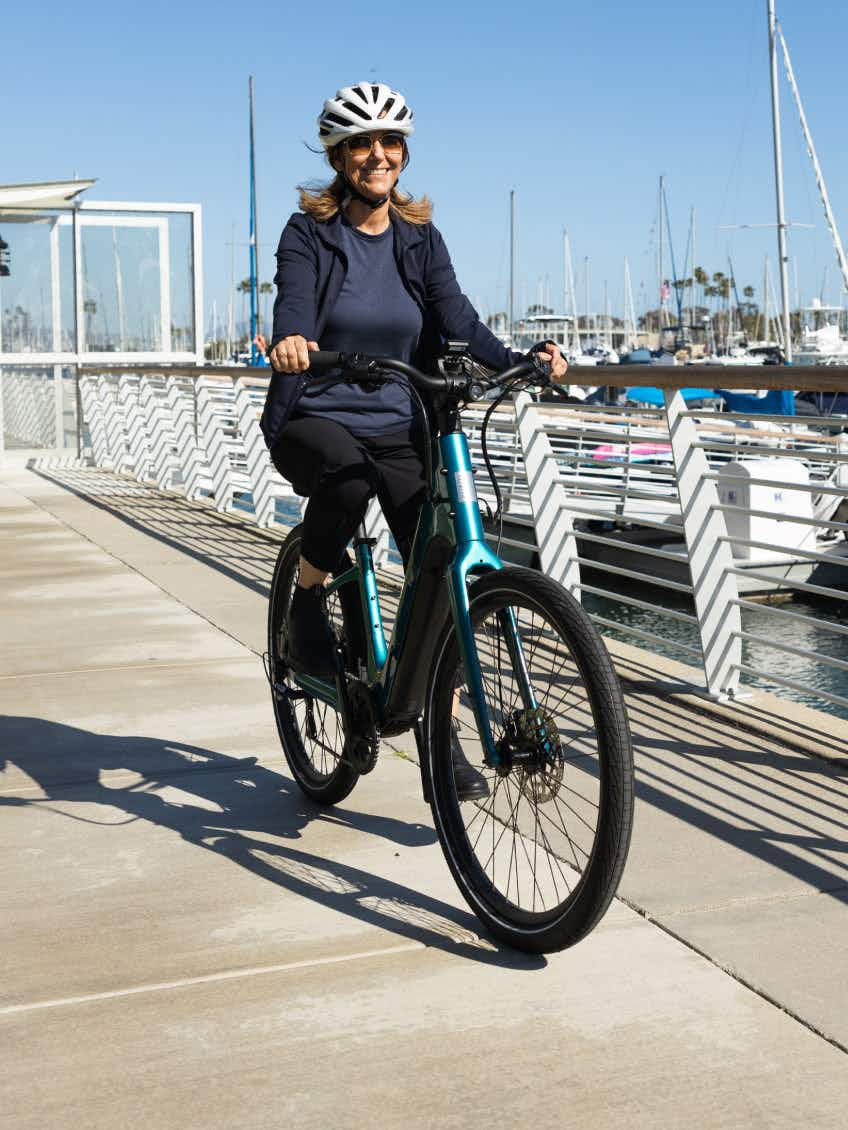 A rider in a helmet and dark clothes cruises along a waterfront path beside docked boats under clear skies on the Aventon Pace 5 REC Step-Through electric bike by Aventon.