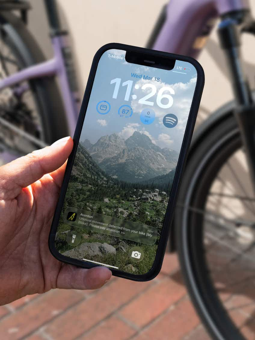 A hand holds a smartphone showing cycling stats and a bike movement alert, with an Aventon Level 4 ADV commuter eBike in the scenic mountain background.