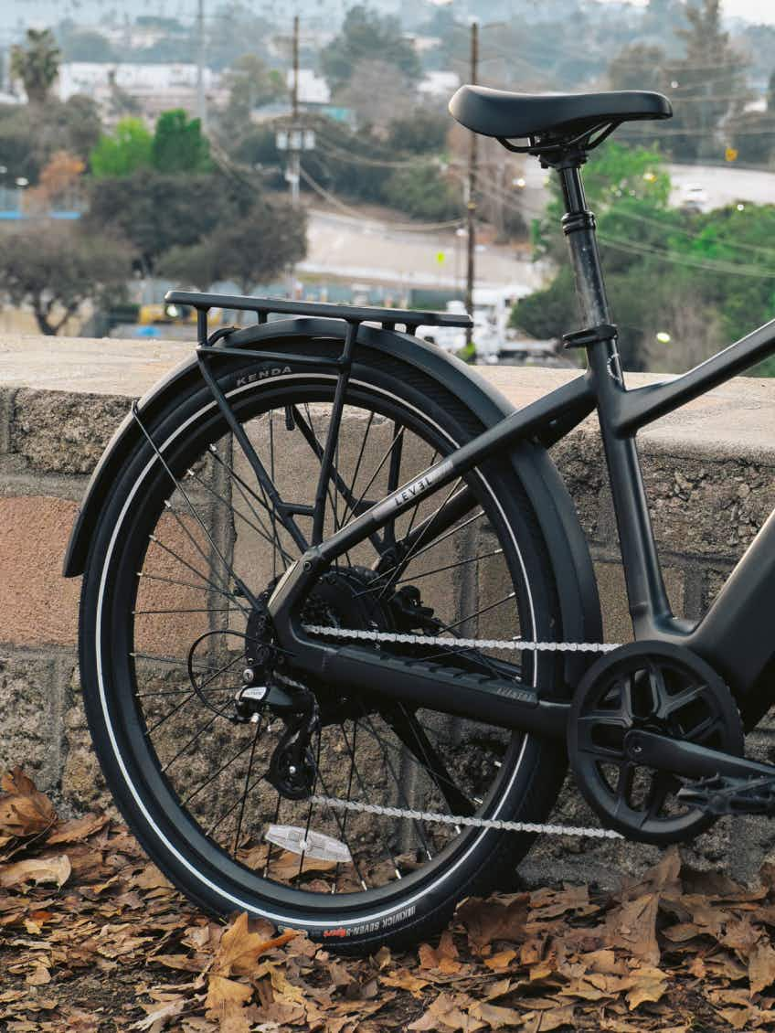 Aventon Level 4 REC by Aventon parked against a stone wall, fallen leaves beneath and cityscape blurred in the background, highlights its powerful hub motor.