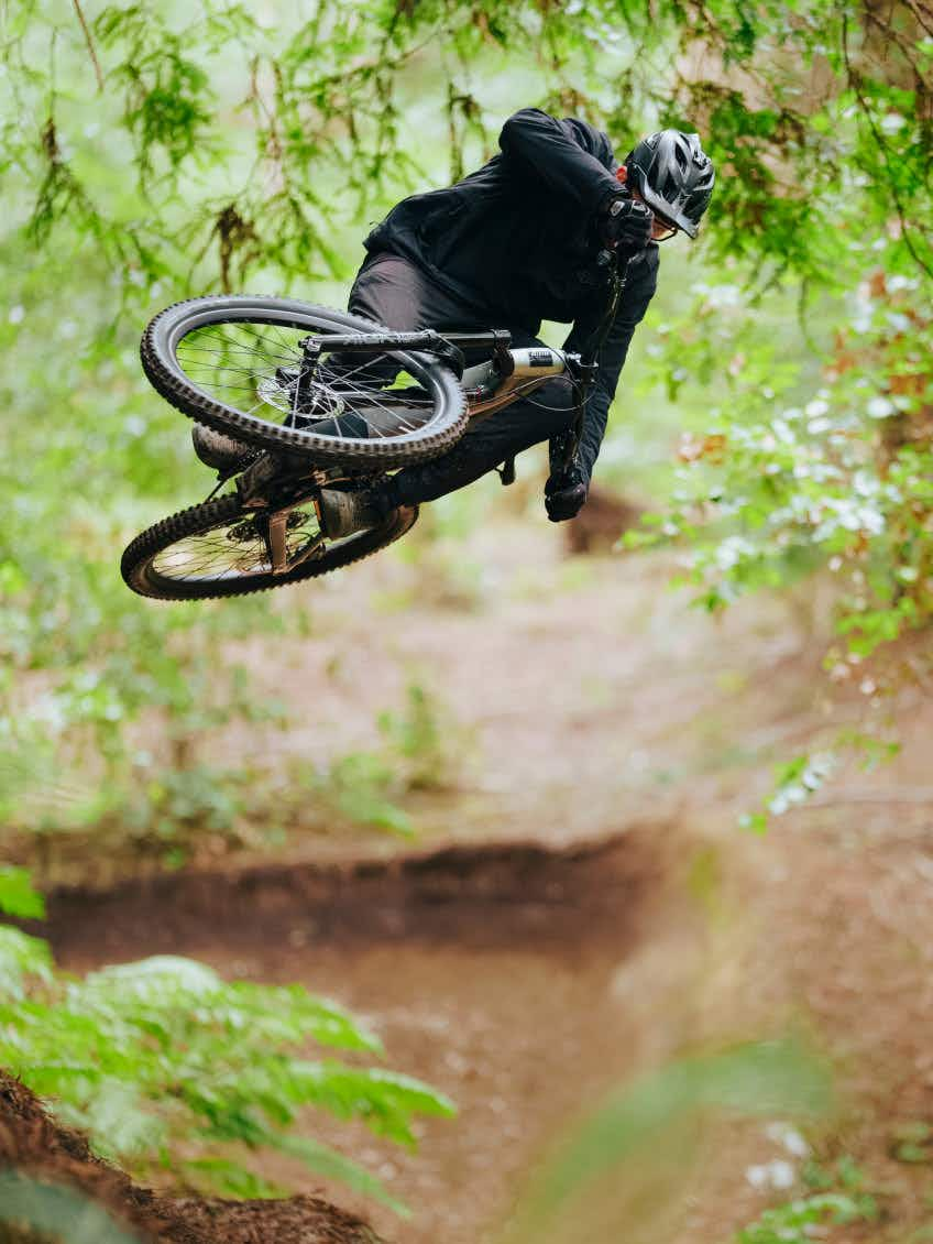 A cyclist in a helmet and dark clothing jumps on an Aventon Current EXP, a carbon electric mountain bike by Aventon with a mid-drive motor, in a forested area.