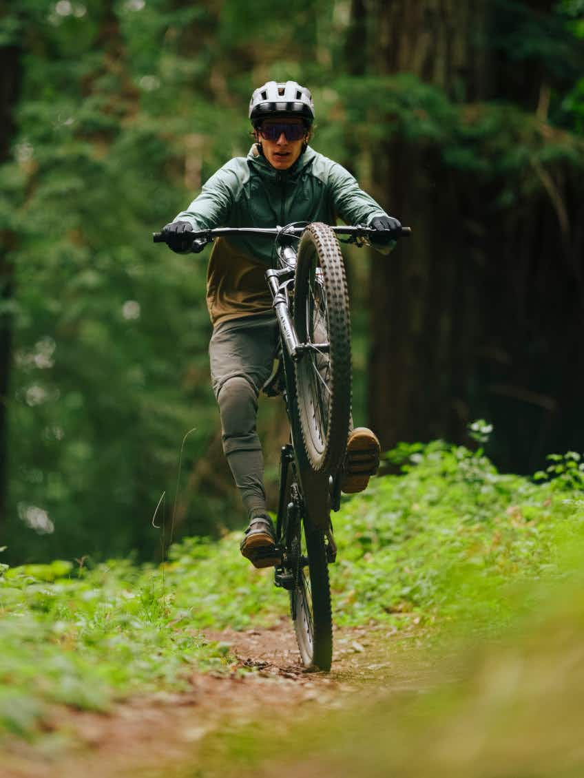 Wearing a helmet and sunglasses, a rider performs a wheelie on an Aventon Current ADV electric mountain bike from Aventon while riding along a forest trail.