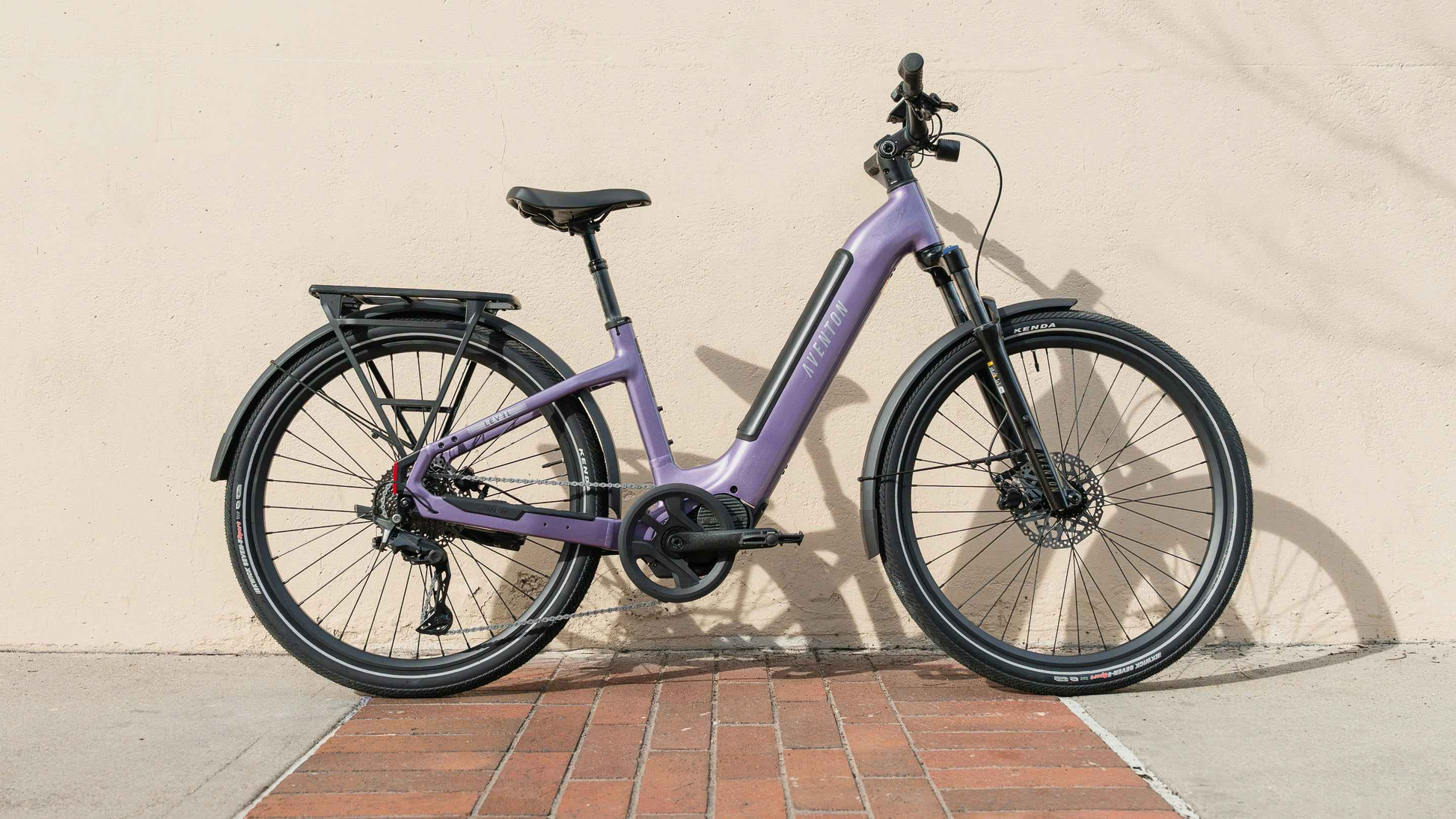 An Aventon Level 4 ADV commuter eBike by Aventon, featuring a purple step-through frame, is parked against a beige wall on a concrete and brick walkway.