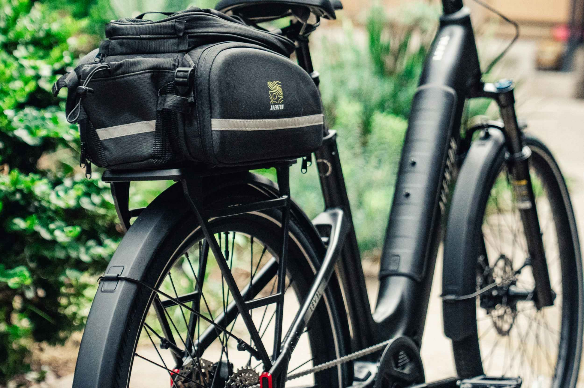 Close-up of the Aventon Level 4 ADV commuter eBike by Aventon, featuring a rear rack with a black pannier bag, parked outdoors among green foliage.