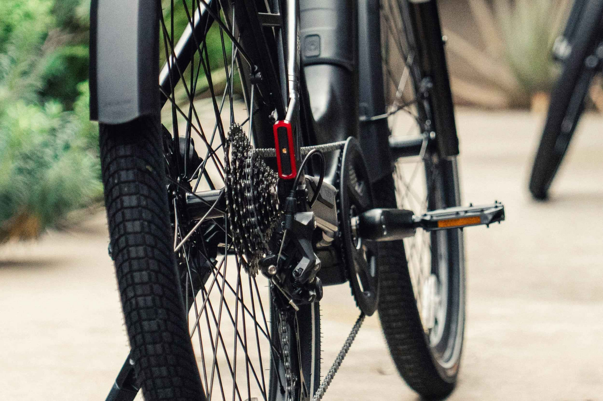 Close-up of the Aventon Level 4 ADV eBike's rear wheel displays its gears, derailleur, chain, and part of the pedal, with a blurred outdoor background. Brand: Aventon.