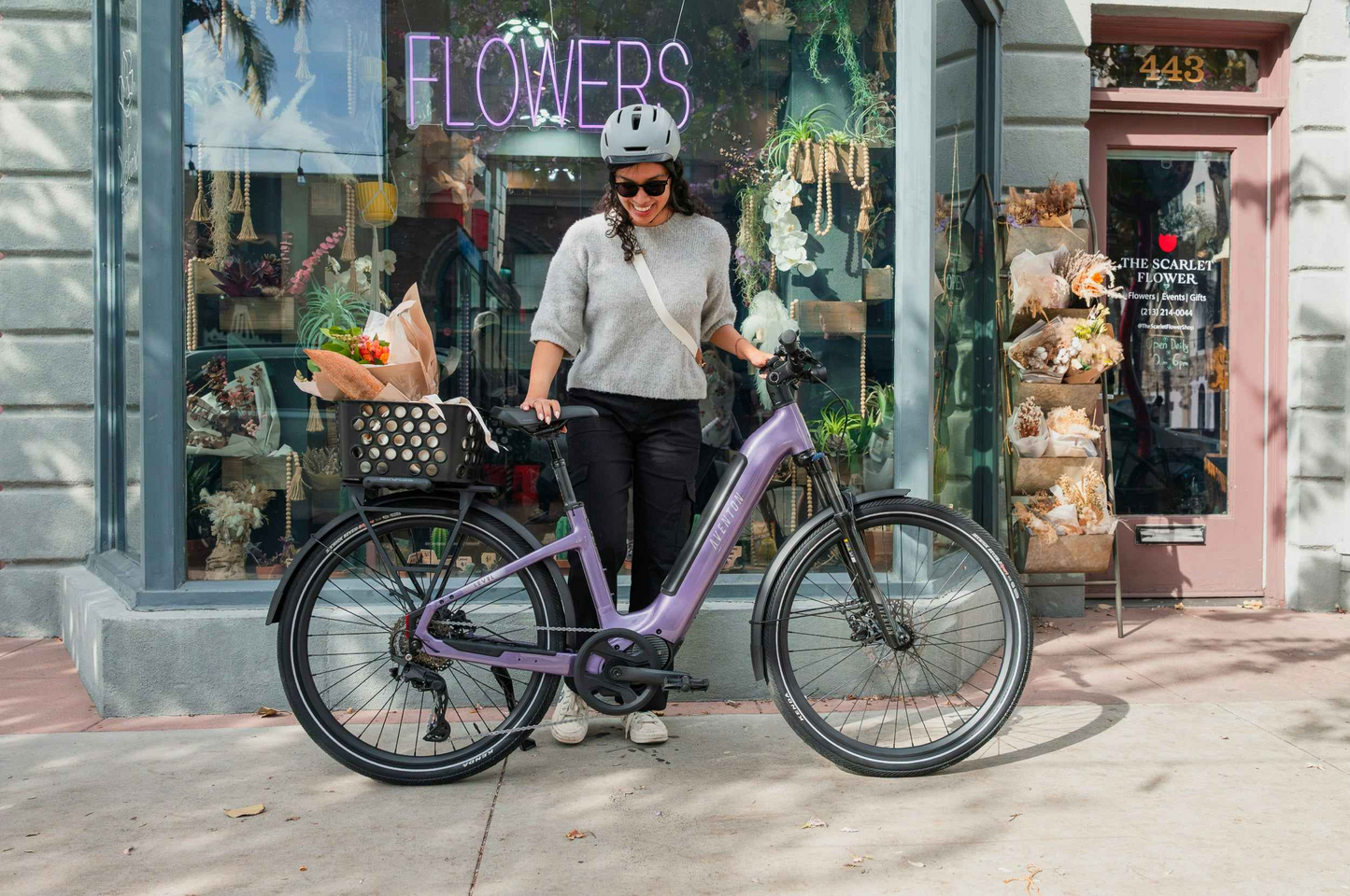 A woman in a helmet stands by her purple Aventon Level 4 ADV eBike from Aventon, its basket brimming with flowers, outside a flower shop.