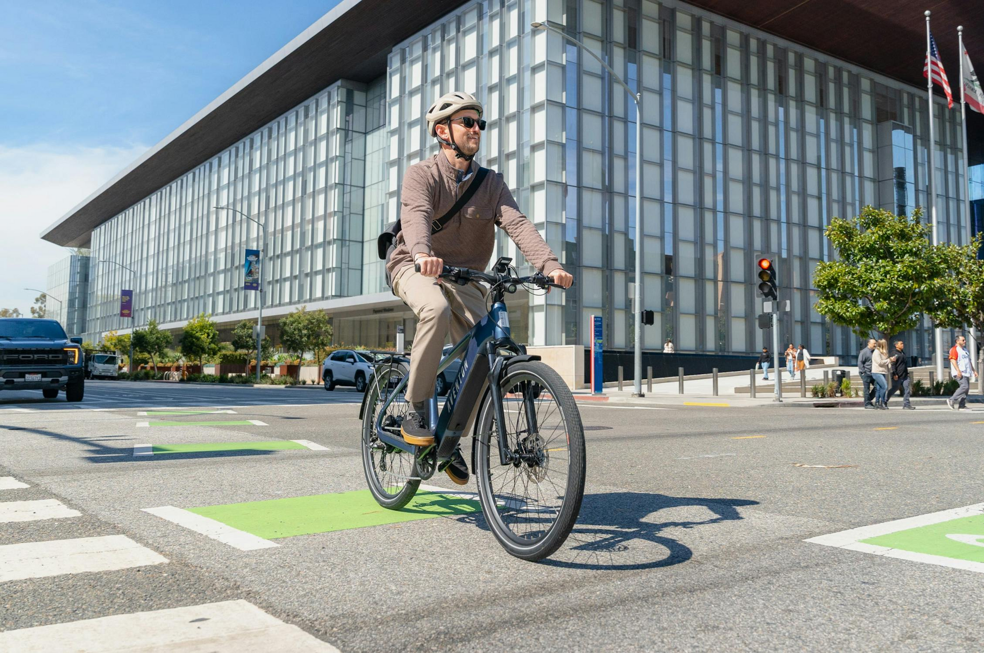 A man in a helmet and sunglasses rides an Aventon Level 4 REC electric bike by Aventon, featuring a powerful hub motor, through a city intersection with modern buildings and traffic lights.