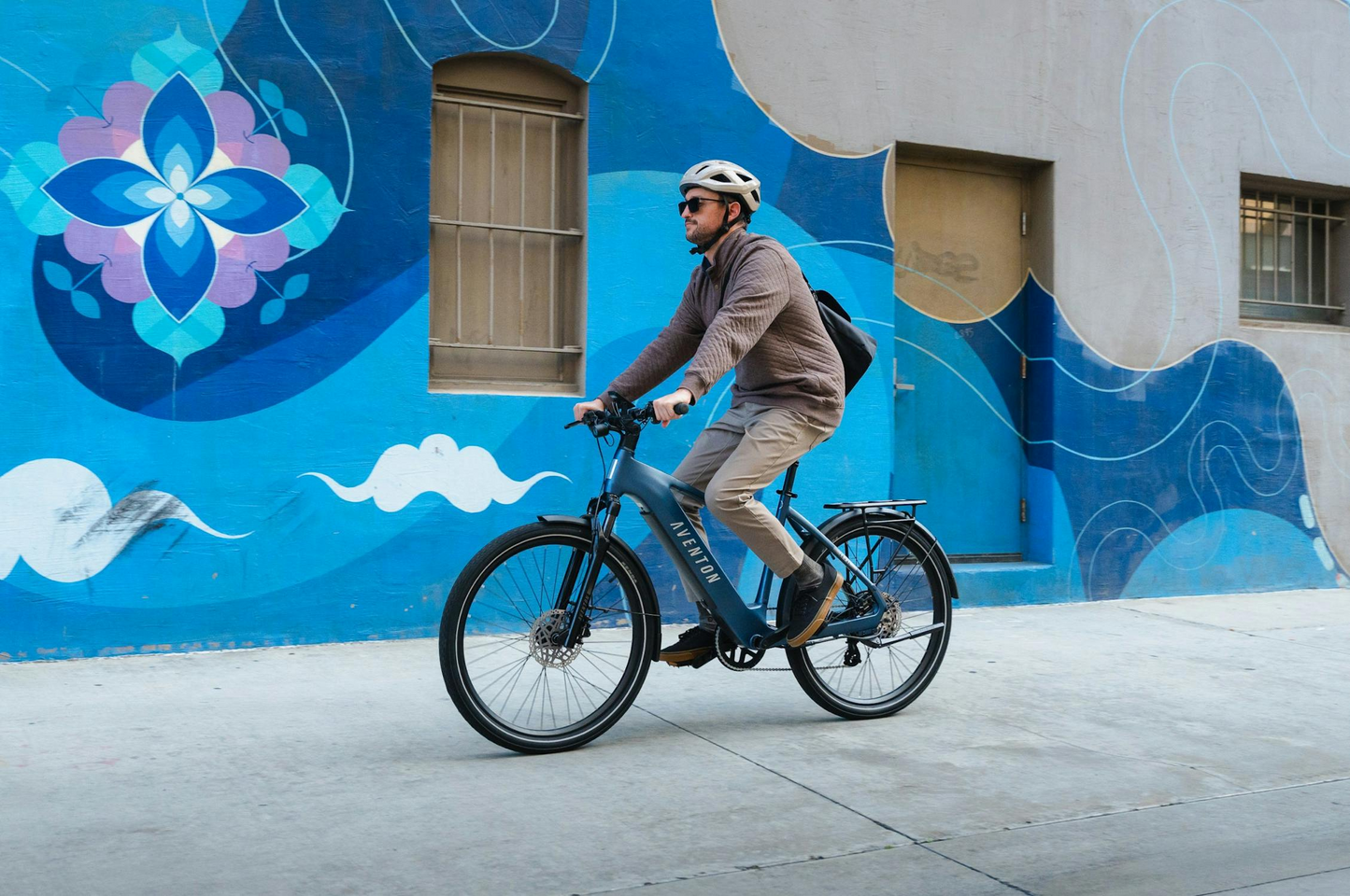 A helmeted rider in sunglasses pedals an Aventon Level 4 REC e-bike by Aventon, featuring a powerful hub motor, past a building with a blue and white mural.