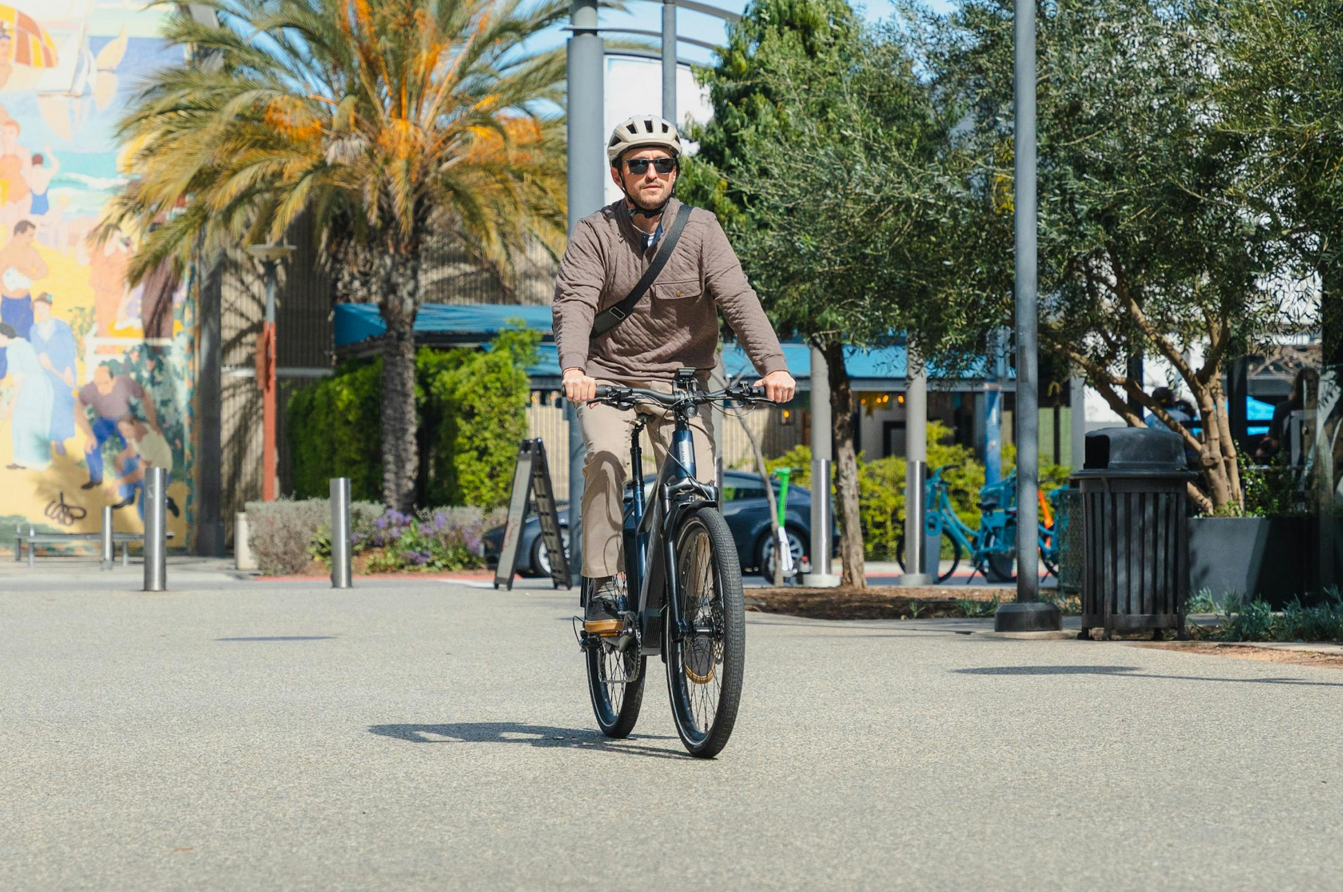 A man in a helmet and sunglasses rides the Aventon Level 4 REC electric bike by Aventon with a powerful hub motor along a paved path in an urban area bordered by palm trees and greenery.