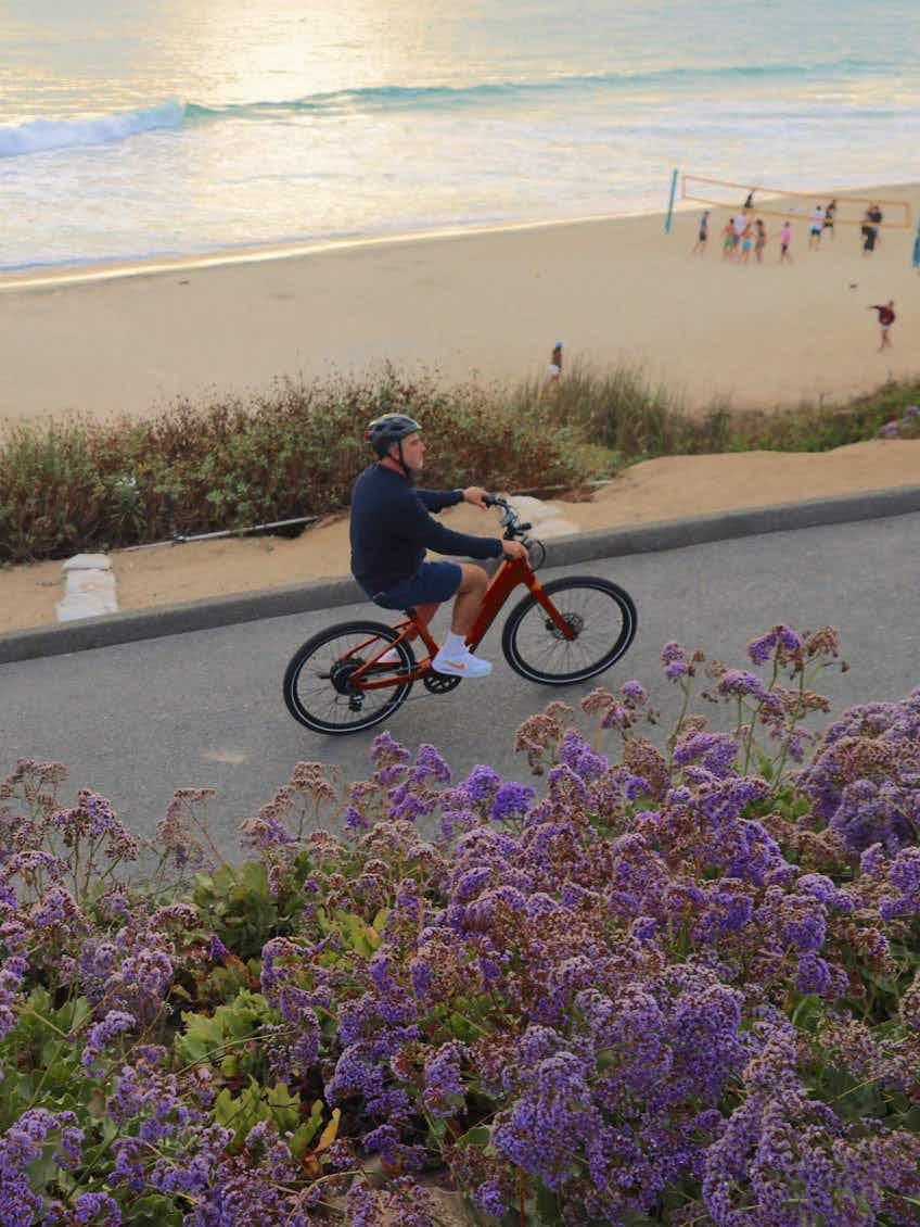 A man wearing a helmet rides an Aventon Pace 5 REC Step-Through by Aventon along a coastal road, with purple flowers in the foreground and people playing volleyball on the beach in the background.