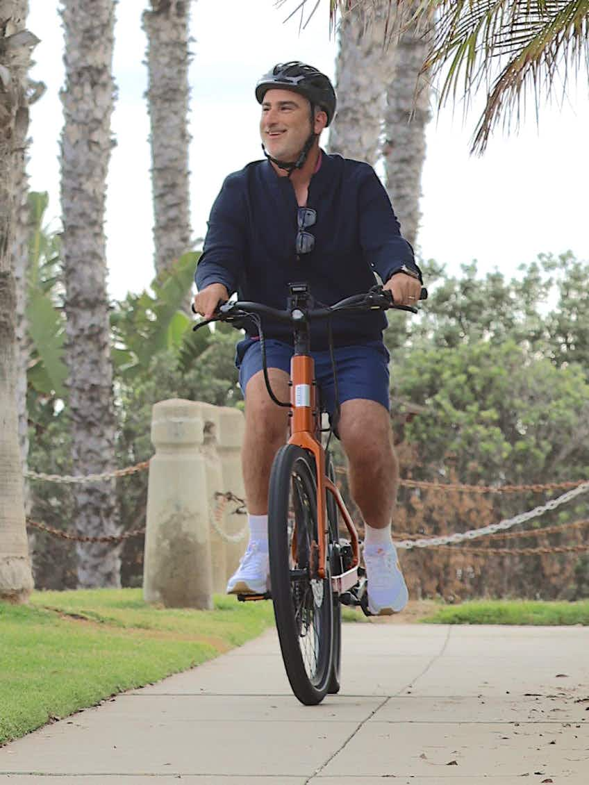 A man in a helmet and casual clothes rides an Aventon Pace 5 REC Step-Through electric bike by Aventon on a tree-lined paved path.