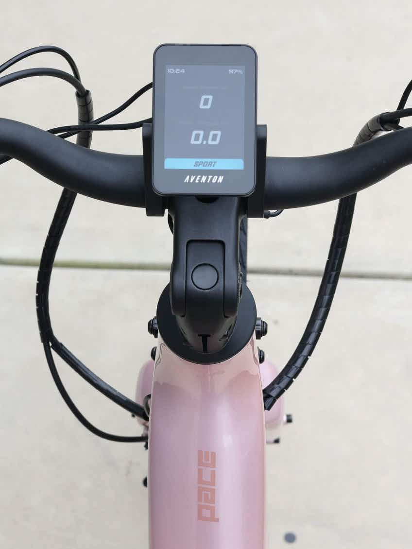 View of the Aventon Pace 5 REC Step-Through electric bike's handlebars and digital display showing speed and battery, set on a pink Aventon comfort cruiser frame against a concrete background.