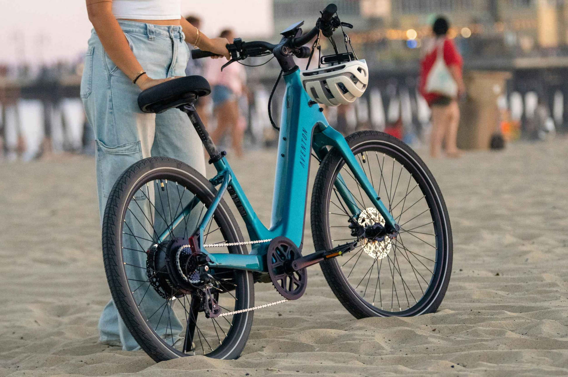 A person in light blue jeans stands on a sandy beach holding an Aventon Pace 5 REC Step-Through electric bike from Aventon, with a white helmet hanging on the handlebars.