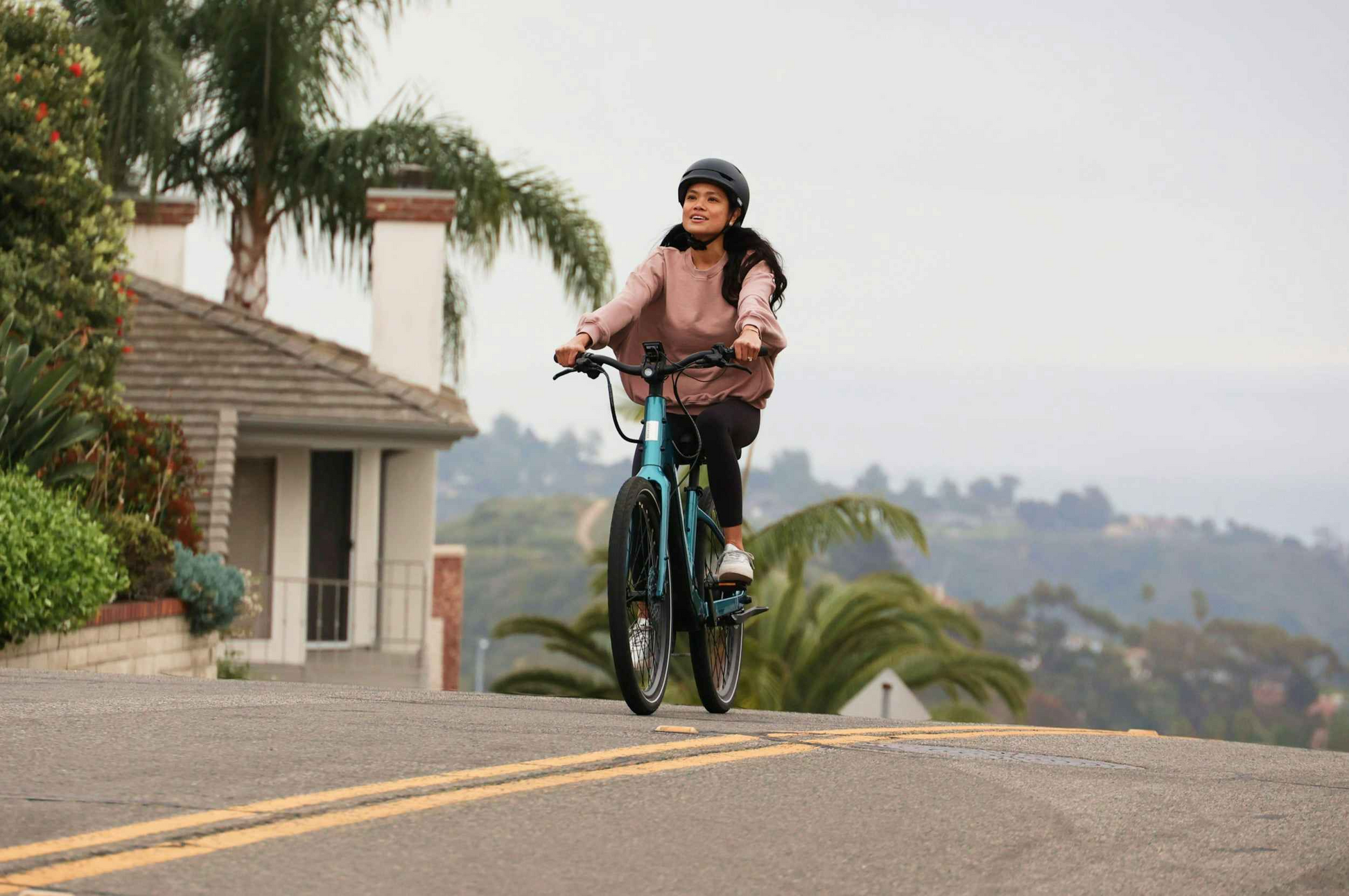 A helmeted rider pedals an Aventon Pace 5 REC Step-Through by Aventon up a suburban road bordered by palm trees and houses.