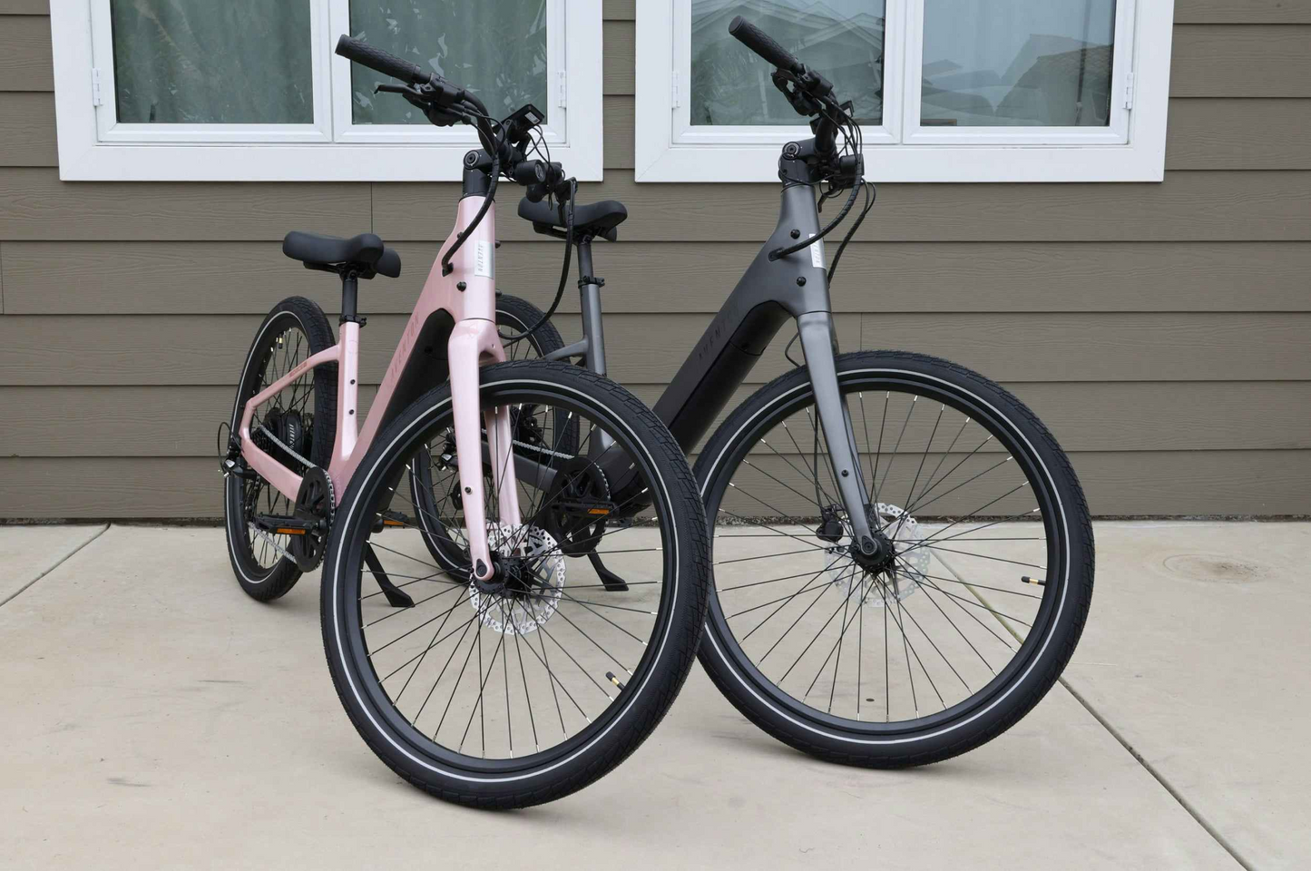 Two modern bikes, including a pink Aventon Pace 5 REC Step-Through electric bike and a black comfort cruiser, are parked on concrete in front of a brown house with white-trimmed windows.